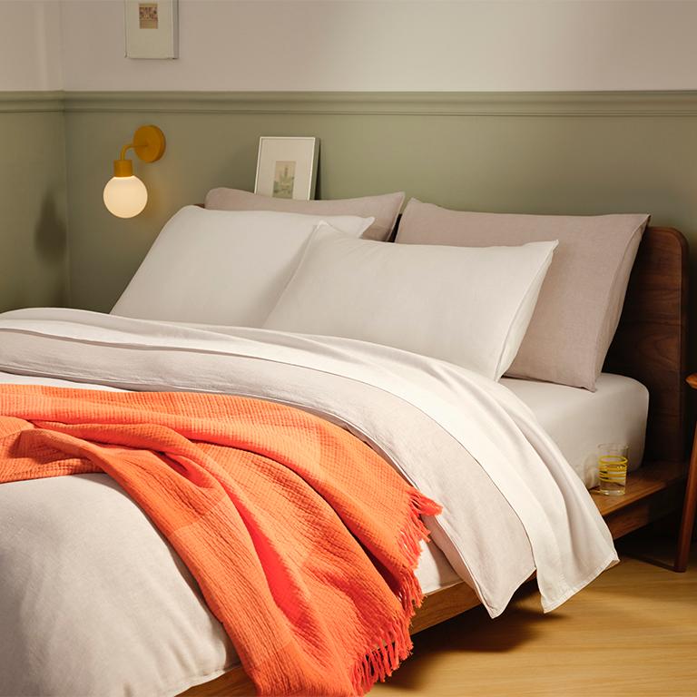 A bed with white and light gray bedding, a vibrant orange throw blanket, a wooden headboard, and two golden wall sconces.