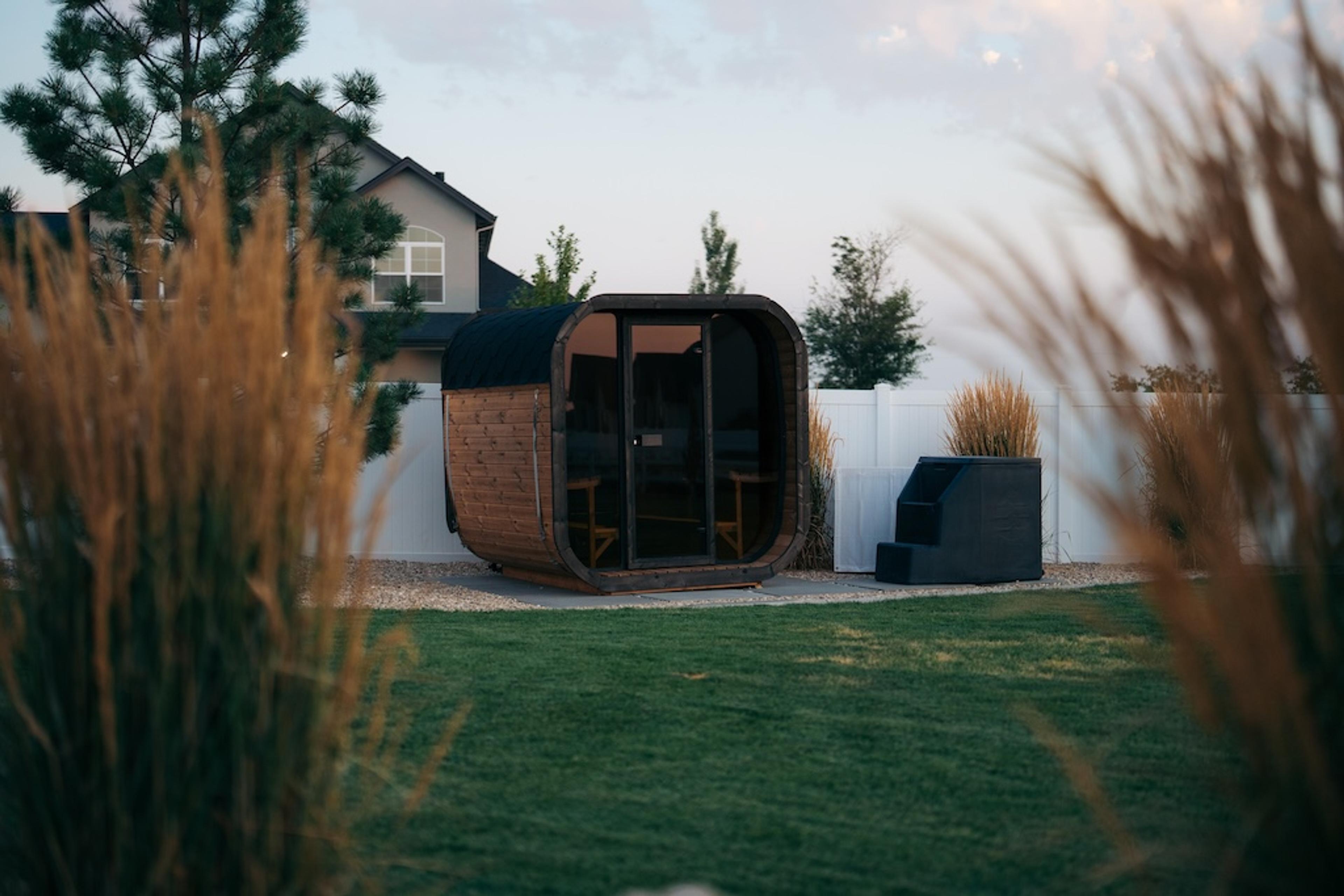 A modern wooden barrel sauna in a backyard with a lawn and a white fence.