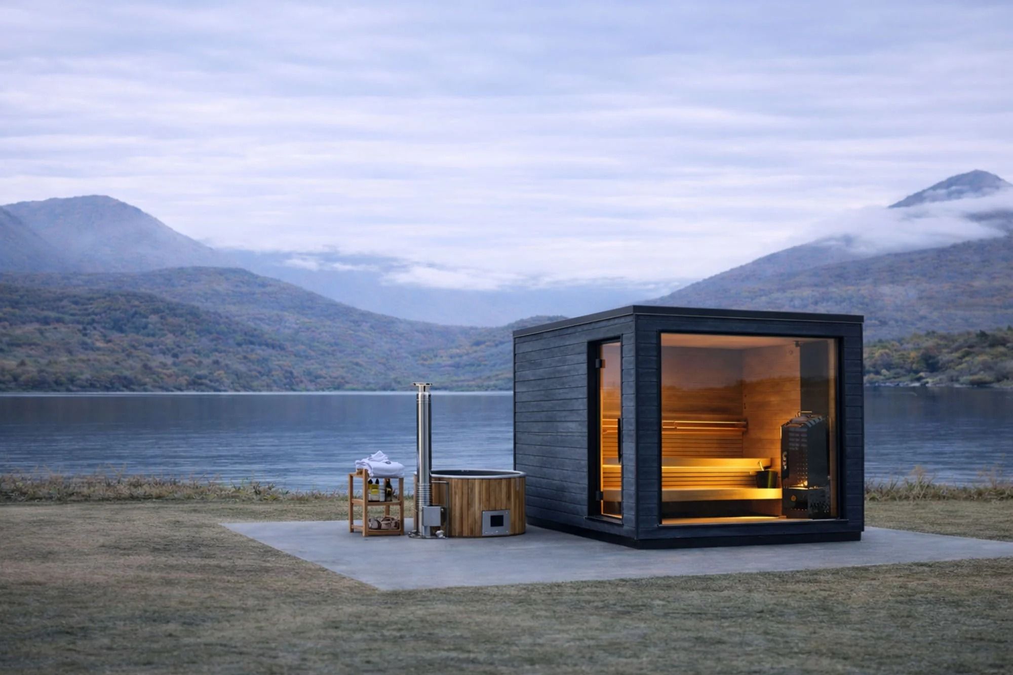 Modern dark sauna with glowing interior and wooden hot tub on a lake shore, backed by misty mountains under an overcast sky.