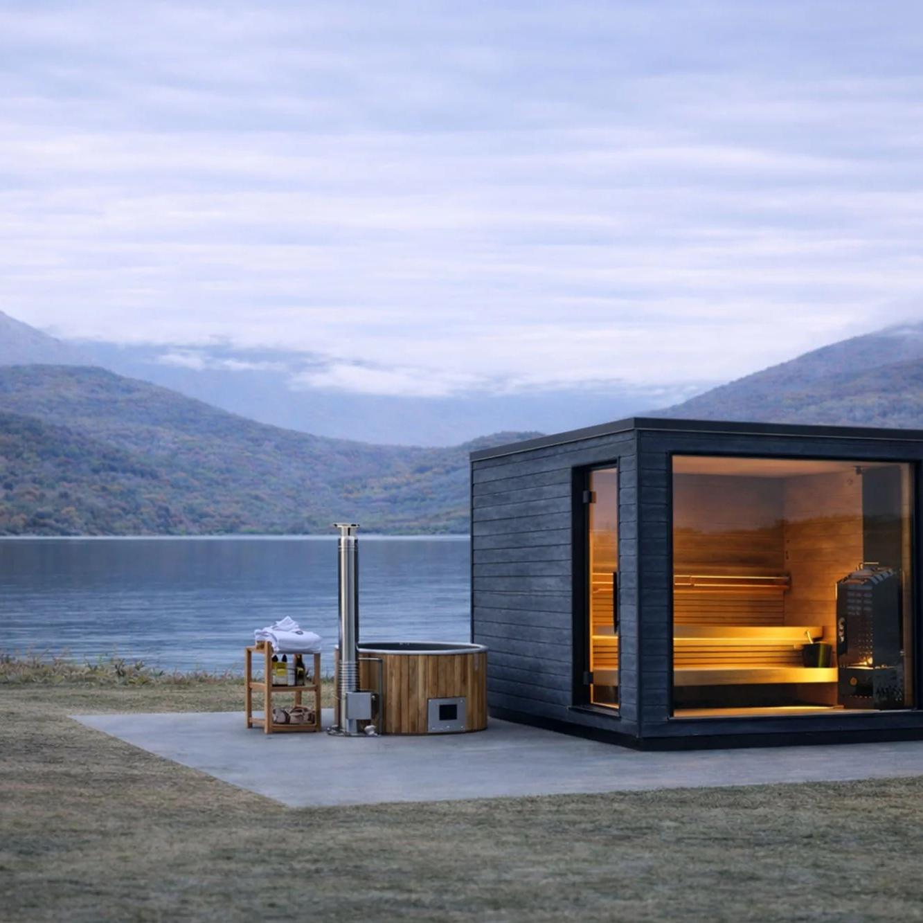 Modern dark sauna with glowing interior and wooden hot tub on a lake shore, backed by misty mountains under an overcast sky.