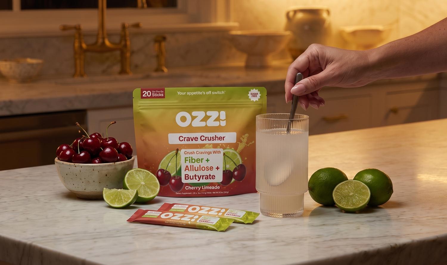 A hand stirs a glass of OZZ! Crave Crusher Cherry Limeade, surrounded by product packaging, fresh cherries, and limes on a kitchen counter.