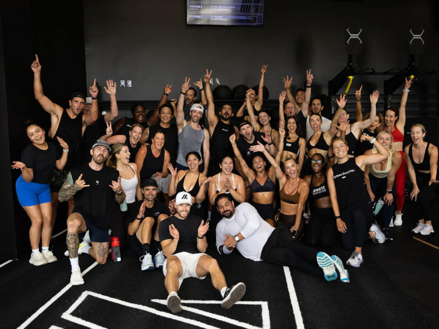 A large, diverse group of smiling people in workout clothes posing excitedly in a gym.