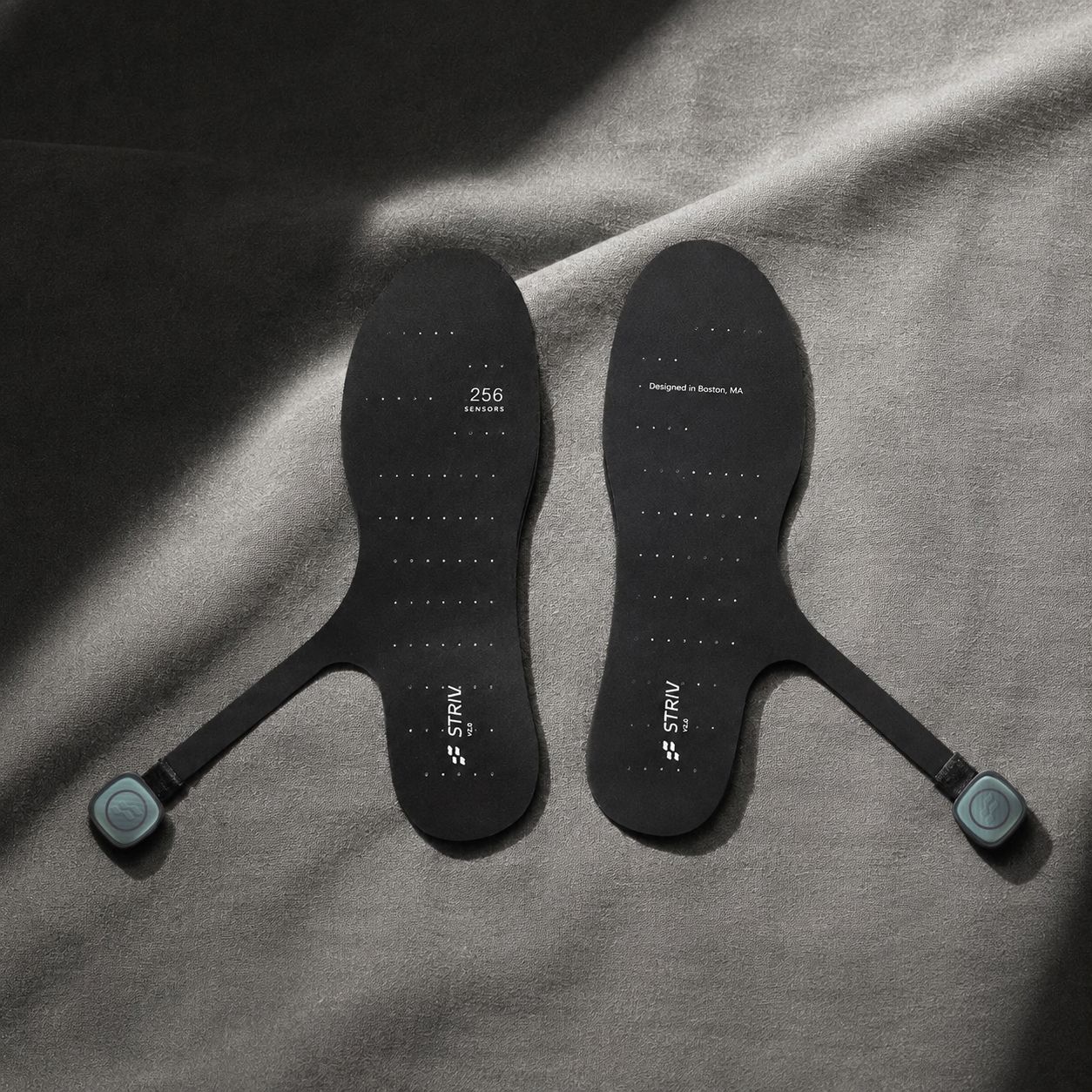 A pair of black smart insoles with teal sensor modules on straps, resting on dark gray fabric.