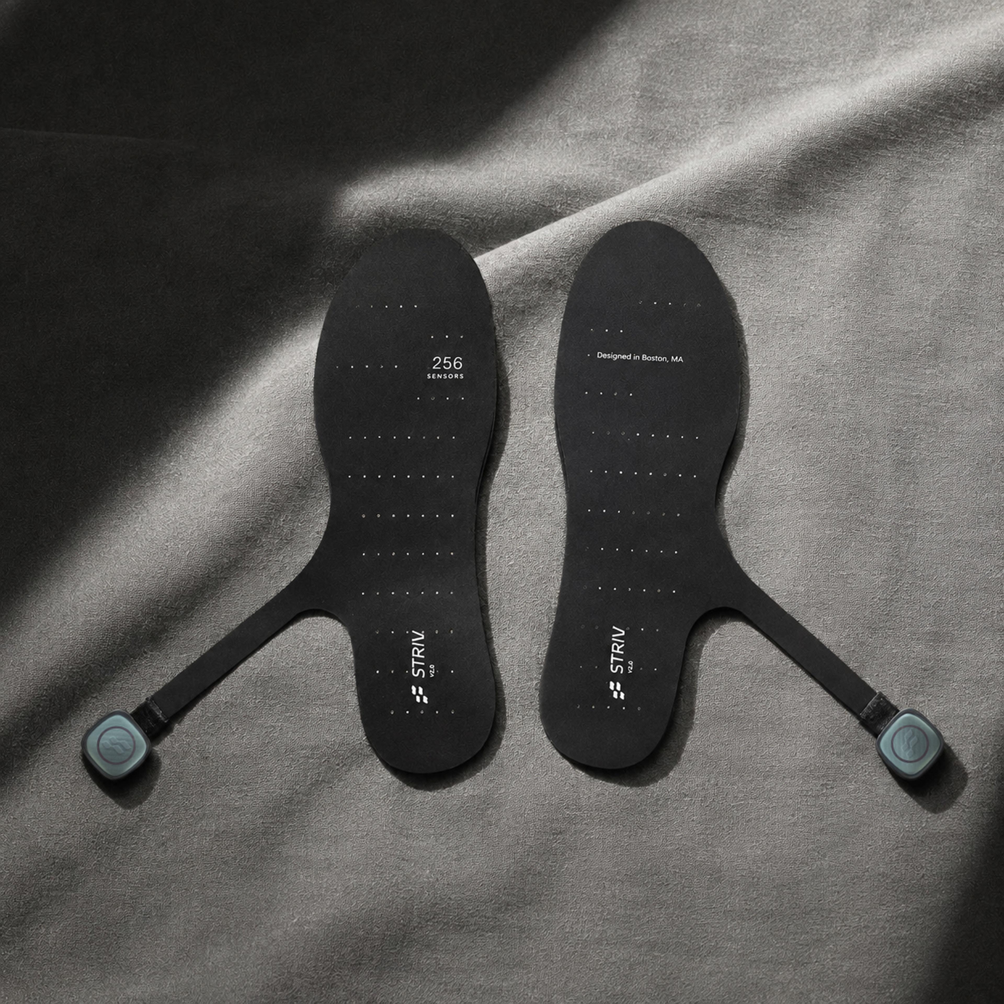 A pair of black smart insoles with teal sensor modules on straps, resting on dark gray fabric.