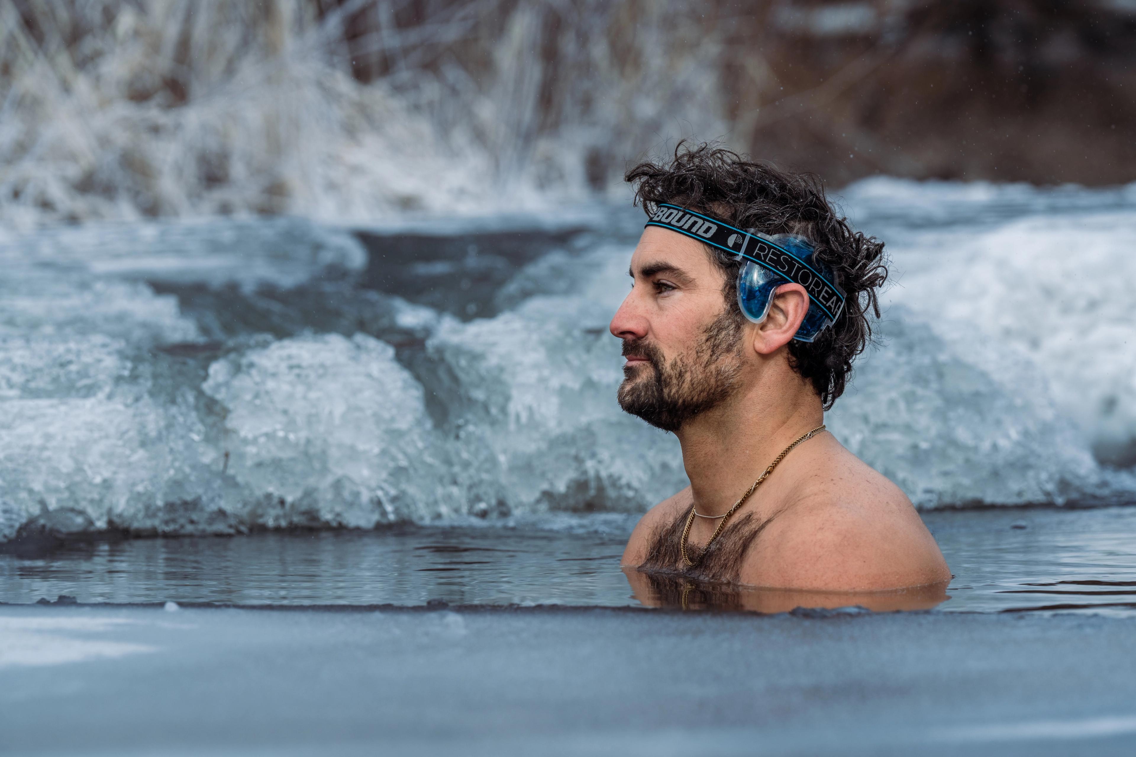 Man with a beard and ear covers submerged in icy water.