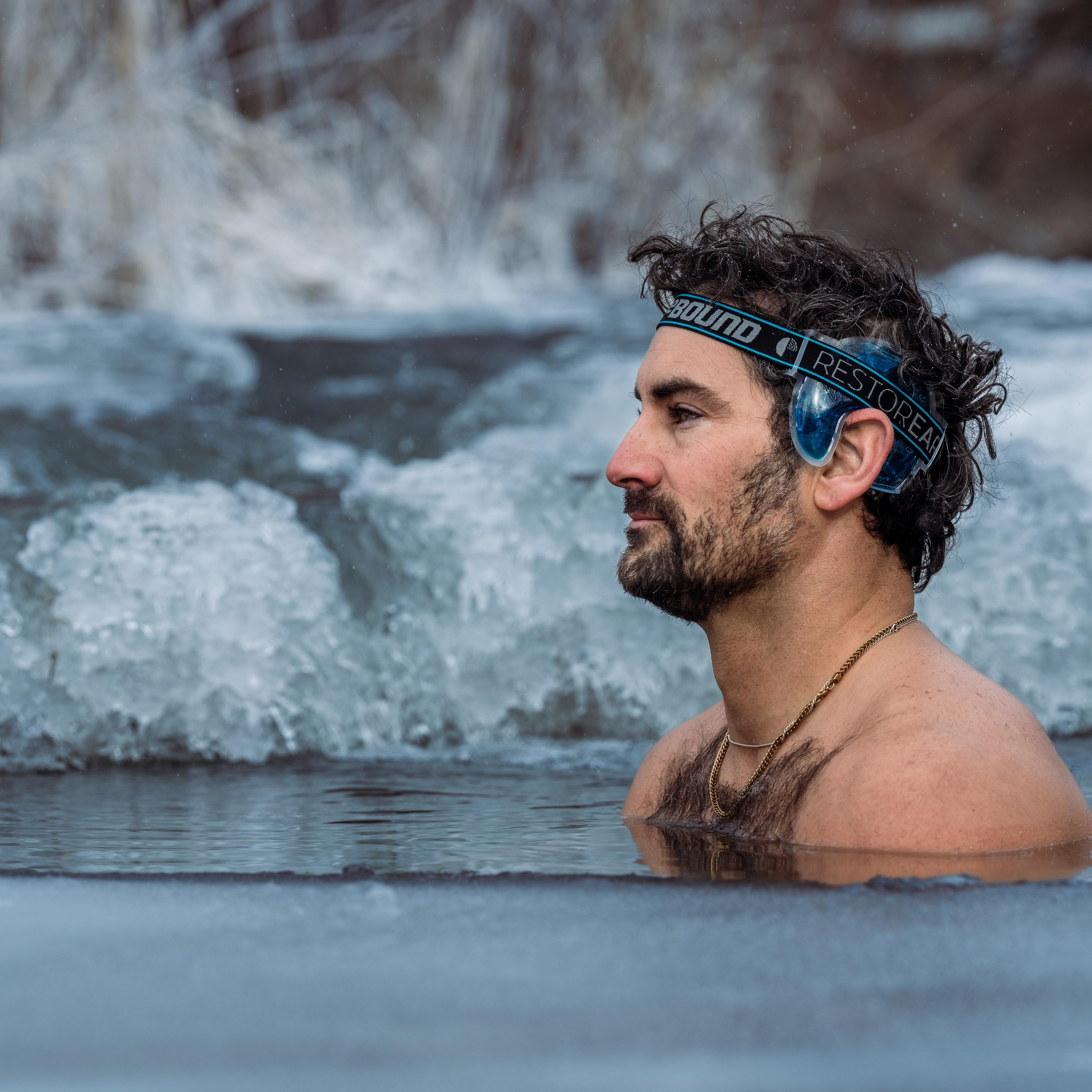 Man with a beard and ear covers submerged in icy water.