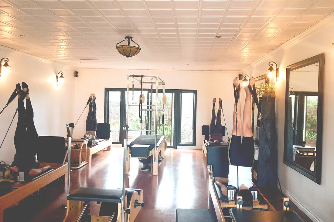 People exercising on Pilates reformers with legs in the air in a brightly lit studio.