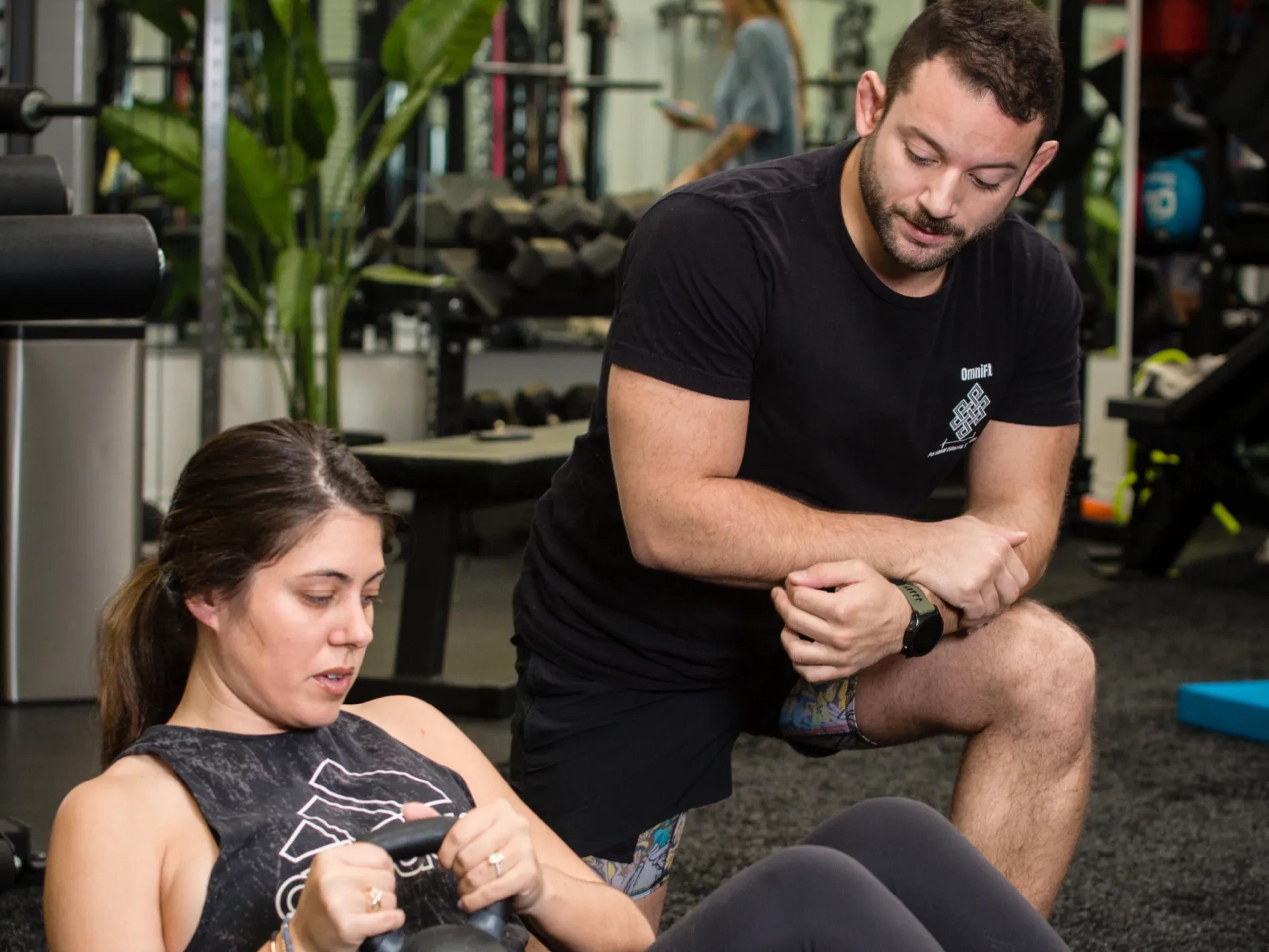 A woman exercising with a kettlebell in a gym, with a male trainer watching.