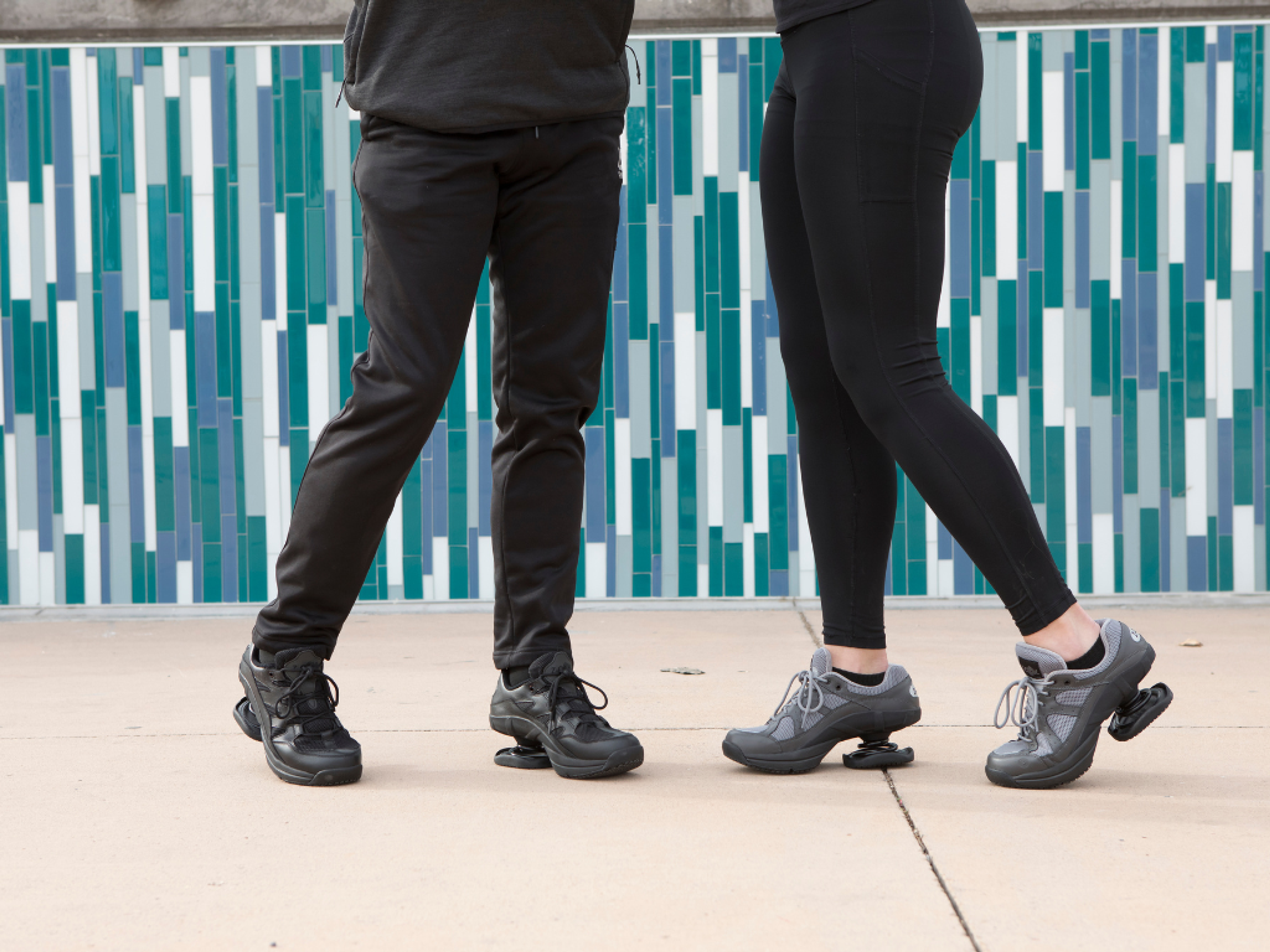 Two people wearing black athletic wear and shoes with spring-loaded soles stand in front of a blue and white tiled wall.