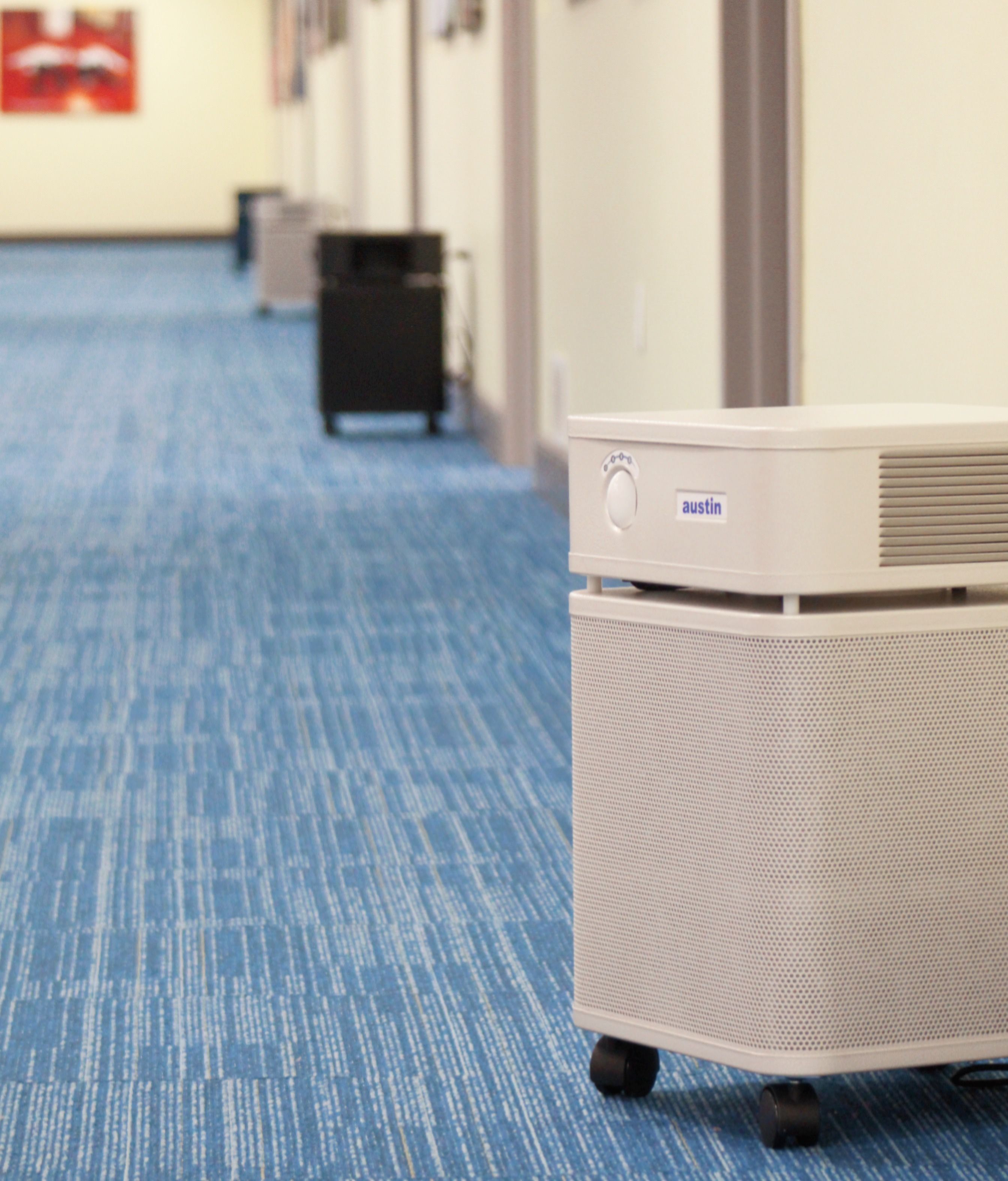 A light gray Austin air purifier on wheels in a blue-carpeted hallway with several other air purifiers visible down the hall.