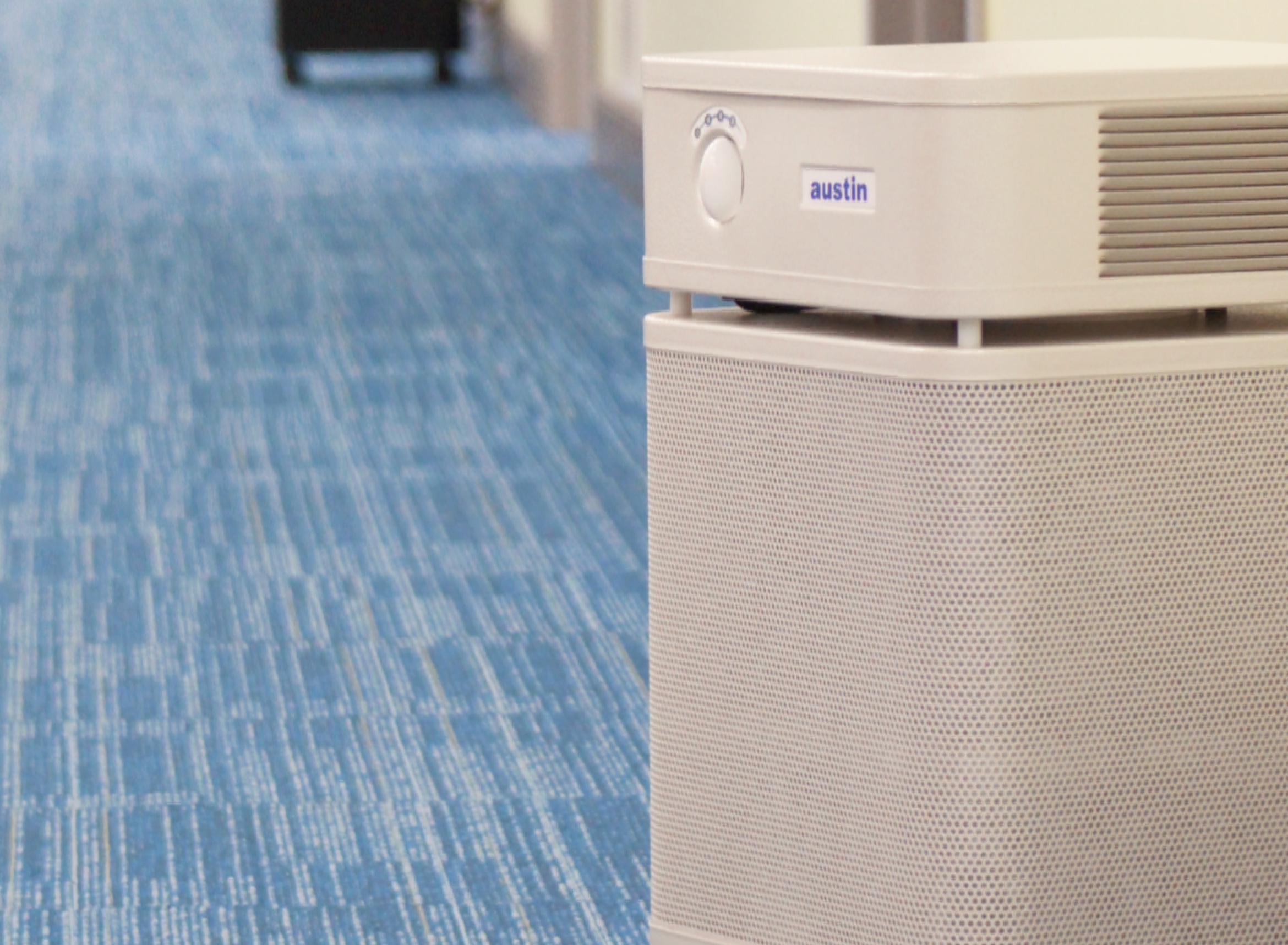 A light gray Austin air purifier on wheels in a blue-carpeted hallway with several other air purifiers visible down the hall.