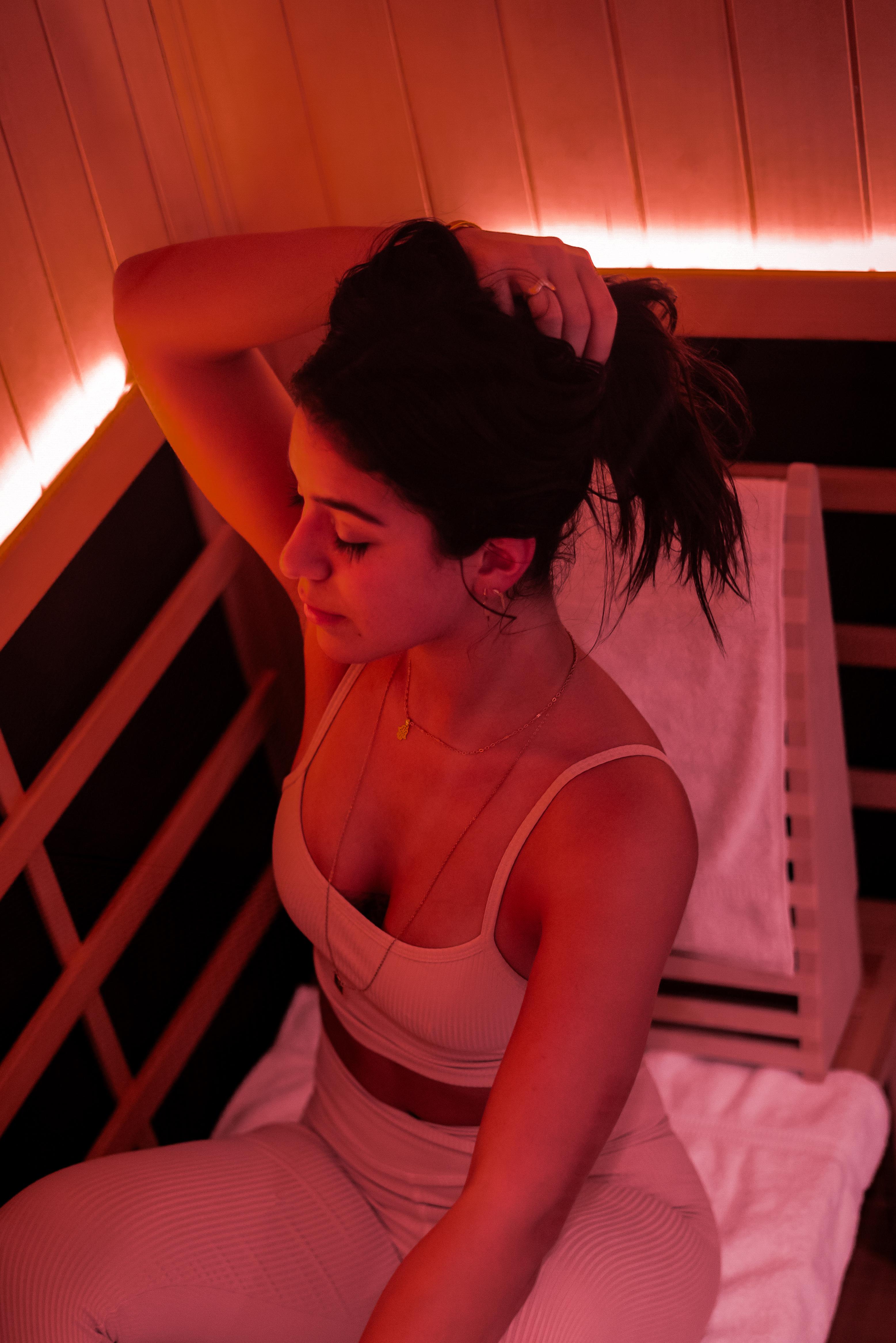 A woman in athletic wear sits with her eyes closed in a red light sauna, adjusting her ponytail.