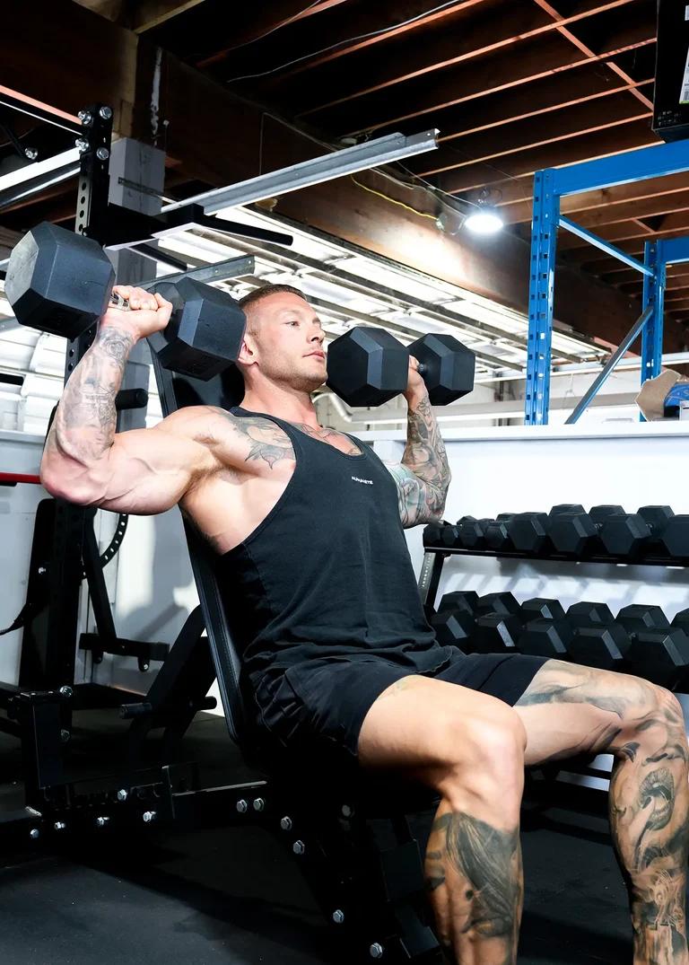 A muscular, tattooed man on an incline bench performs dumbbell shoulder presses in a gym.