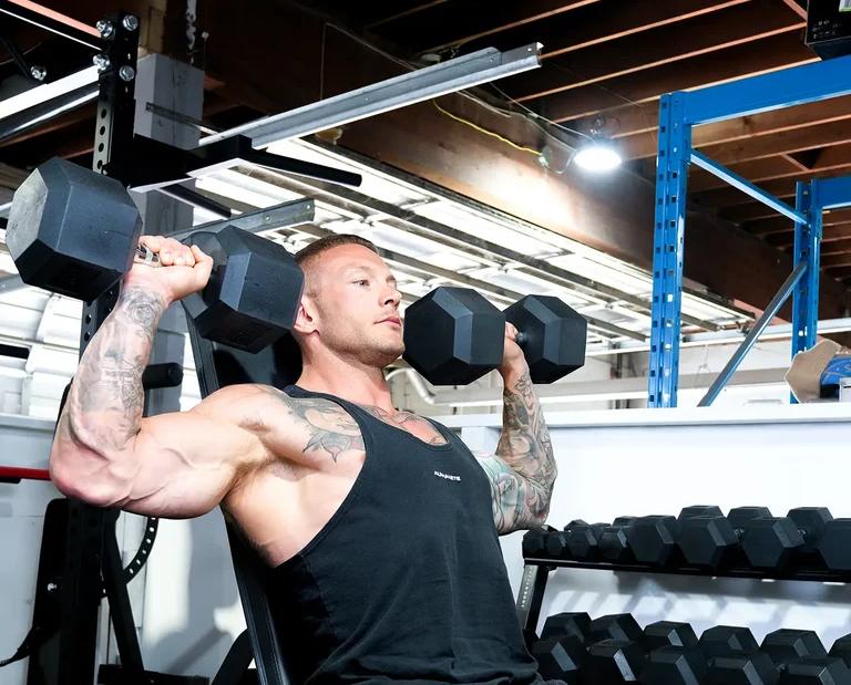 A muscular, tattooed man on an incline bench performs dumbbell shoulder presses in a gym.