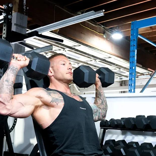 A muscular, tattooed man on an incline bench performs dumbbell shoulder presses in a gym.