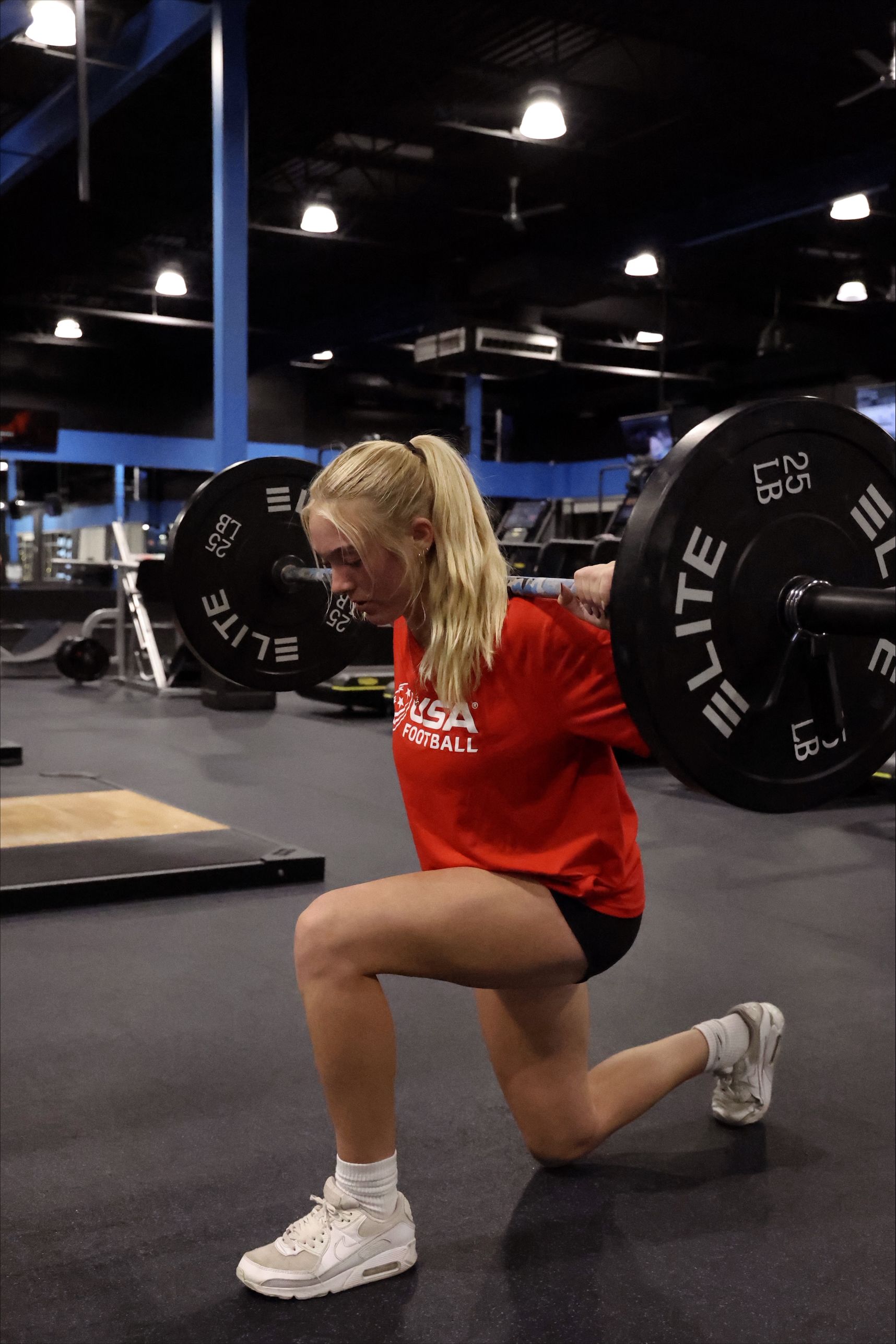 A blonde woman in a red shirt performs a barbell lunge in a gym.
