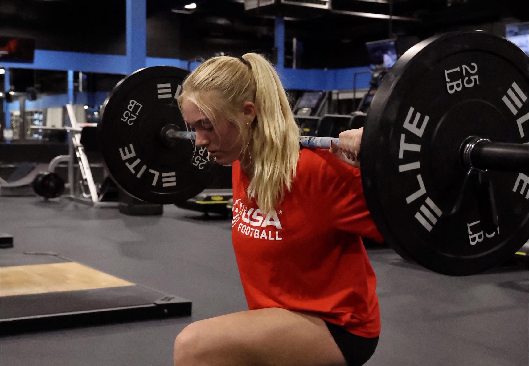A blonde woman in a red shirt performs a barbell lunge in a gym.