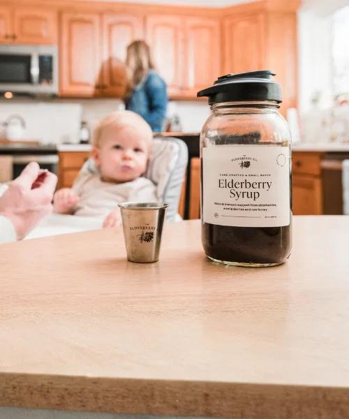 A bottle of Elderberry Syrup and a small cup on a table, with a blurred baby being fed in a kitchen background.