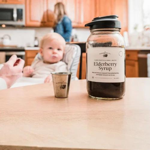 A bottle of Elderberry Syrup and a small cup on a table, with a blurred baby being fed in a kitchen background.