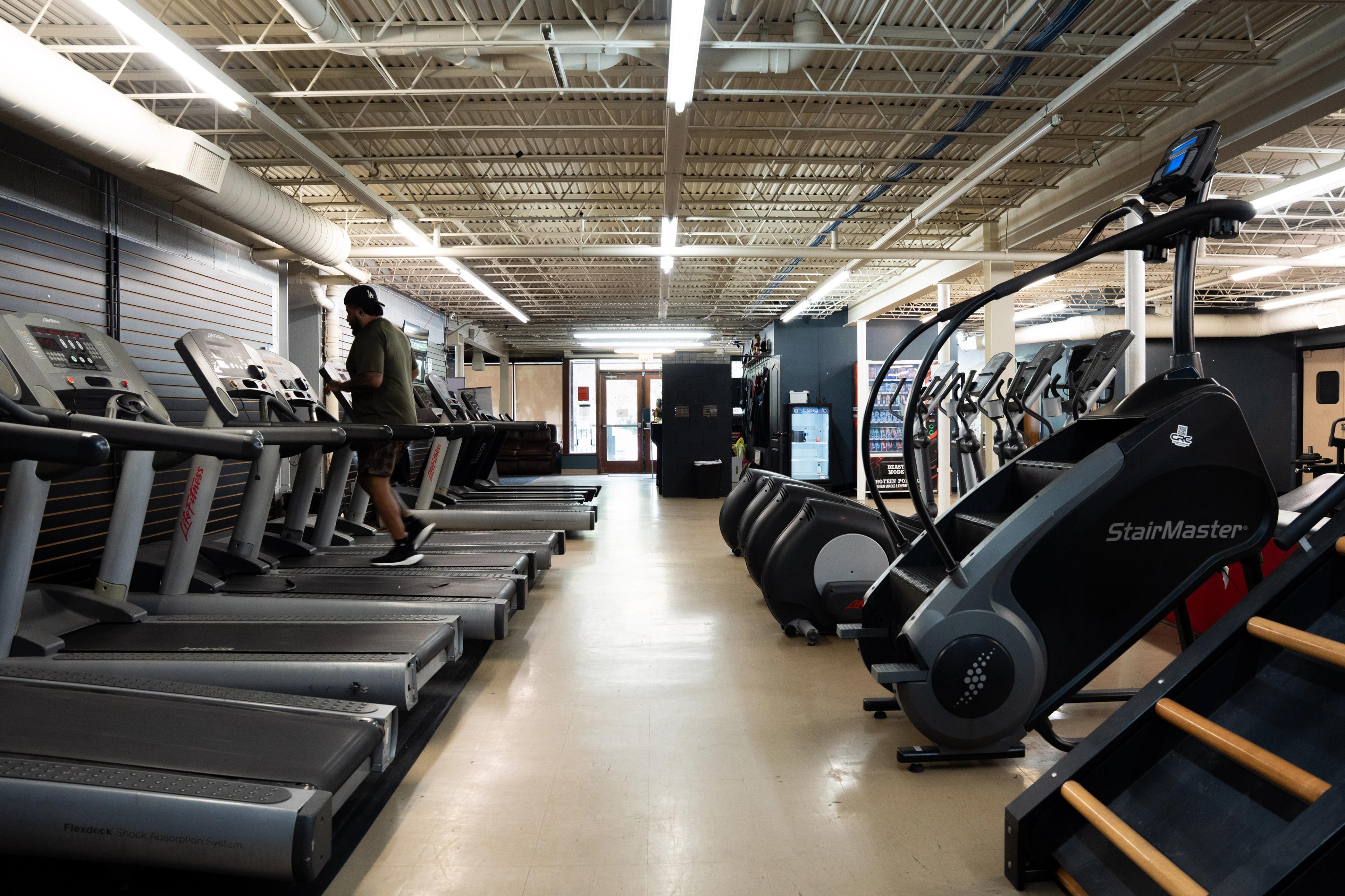 A person on a treadmill in a gym, with rows of treadmills and StairMaster machines.