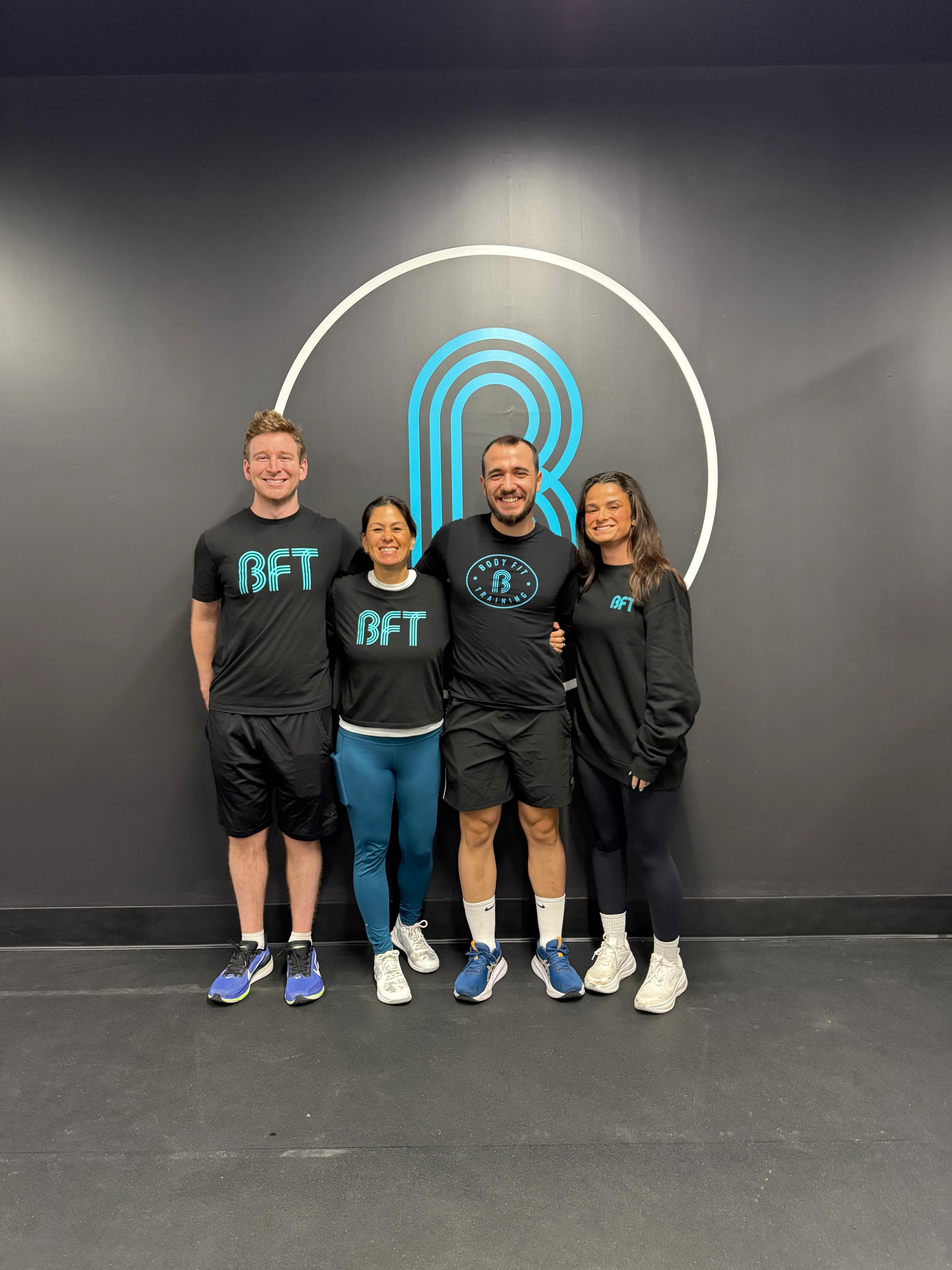 Four smiling people in black BFT athletic wear stand in front of a dark gym wall with a large blue 'B' logo.