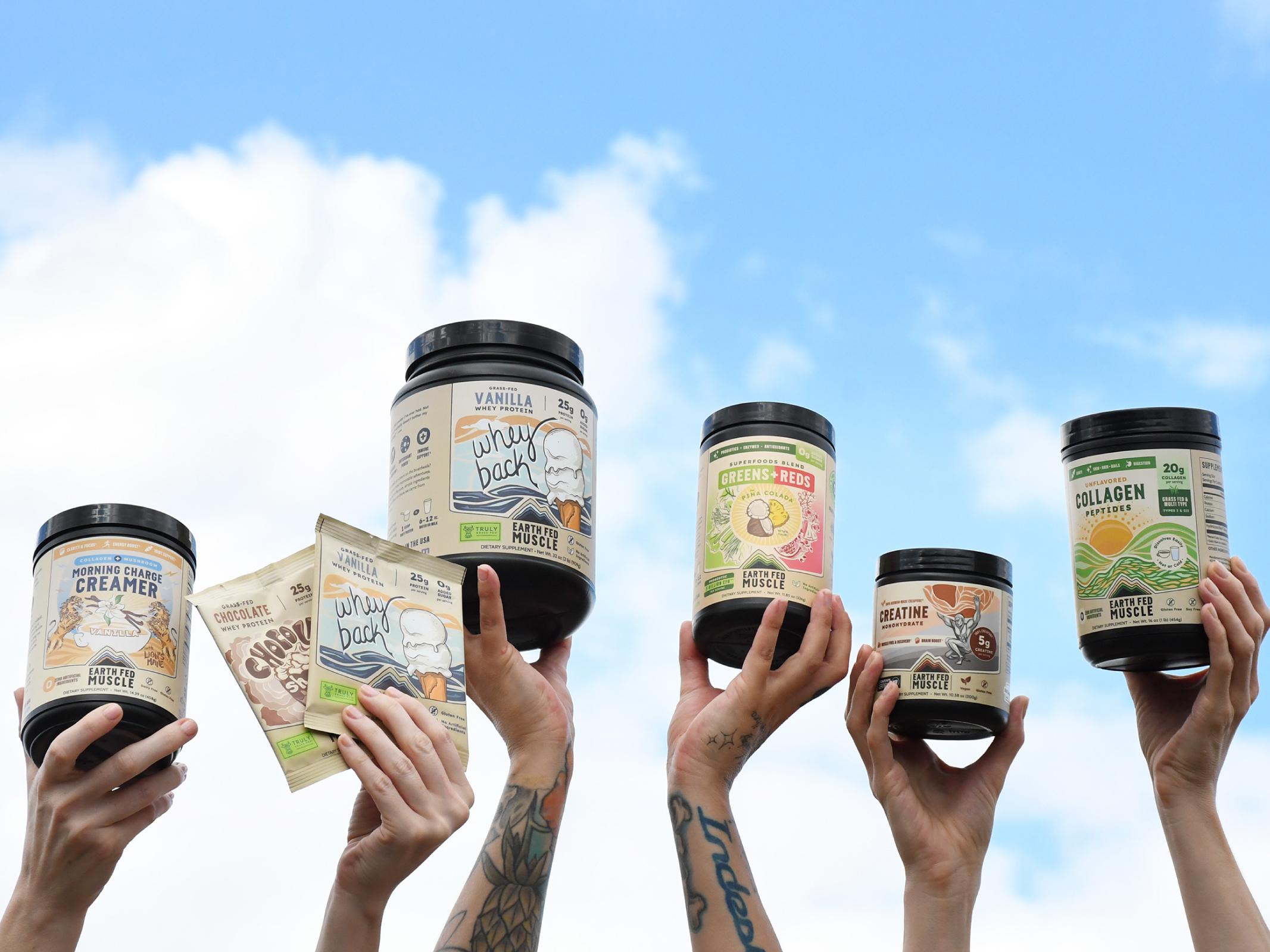 Multiple hands hold various Earth Fed Muscle supplement products against a blue sky with clouds.