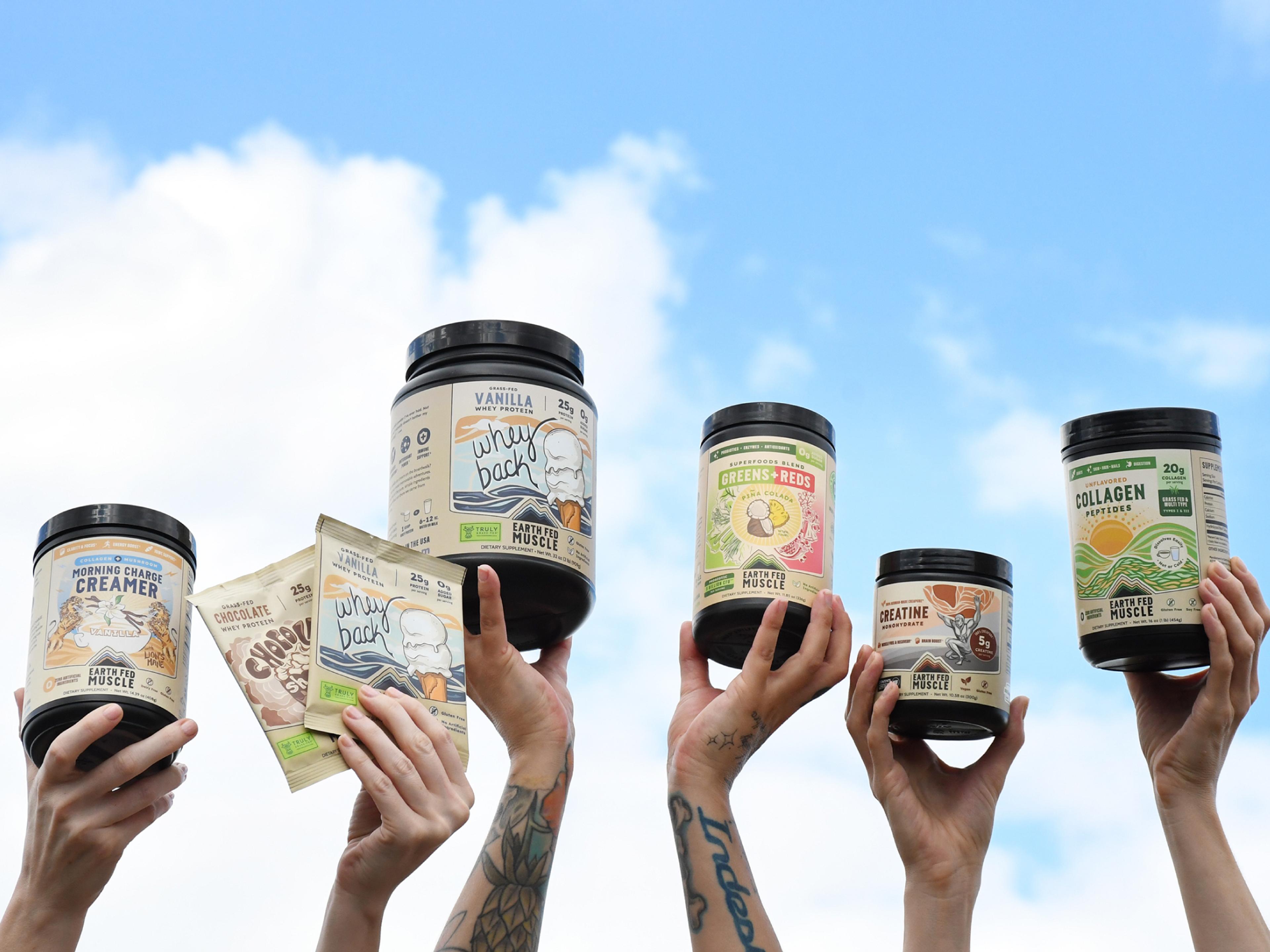 Multiple hands hold various Earth Fed Muscle supplement products against a blue sky with clouds.