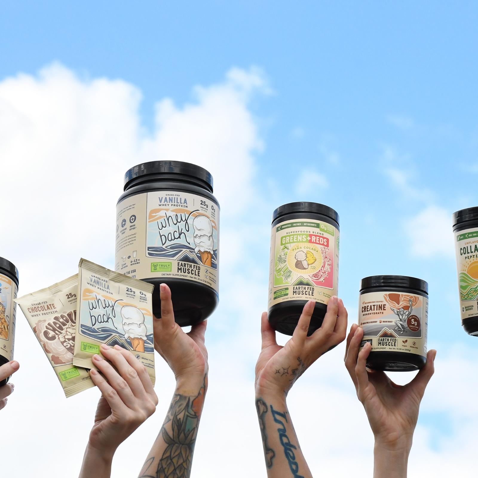 Multiple hands hold various Earth Fed Muscle supplement products against a blue sky with clouds.