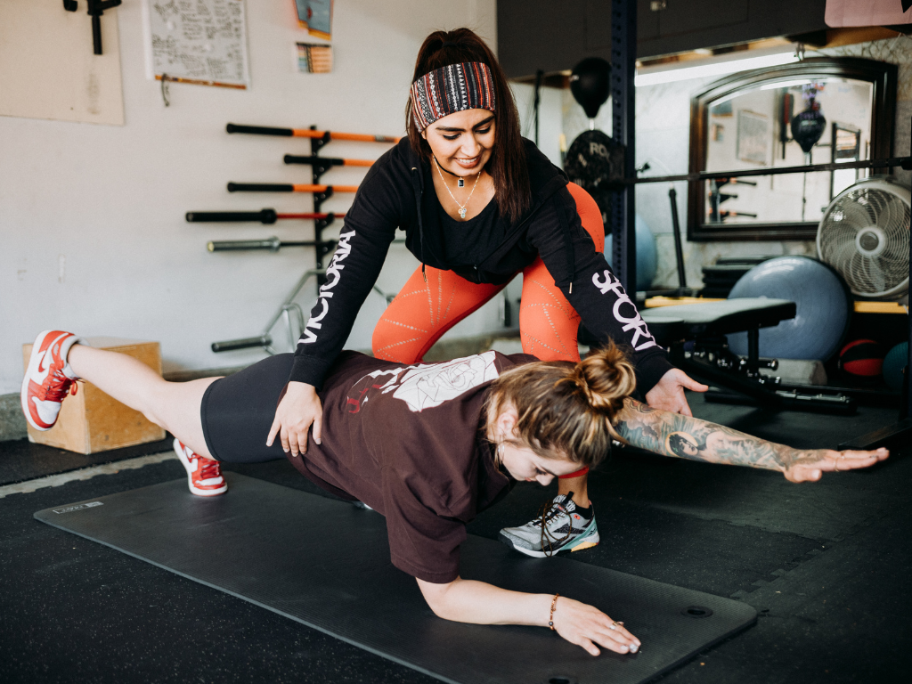 A fitness instructor assists a woman performing a plank with one arm and leg extended in a gym.