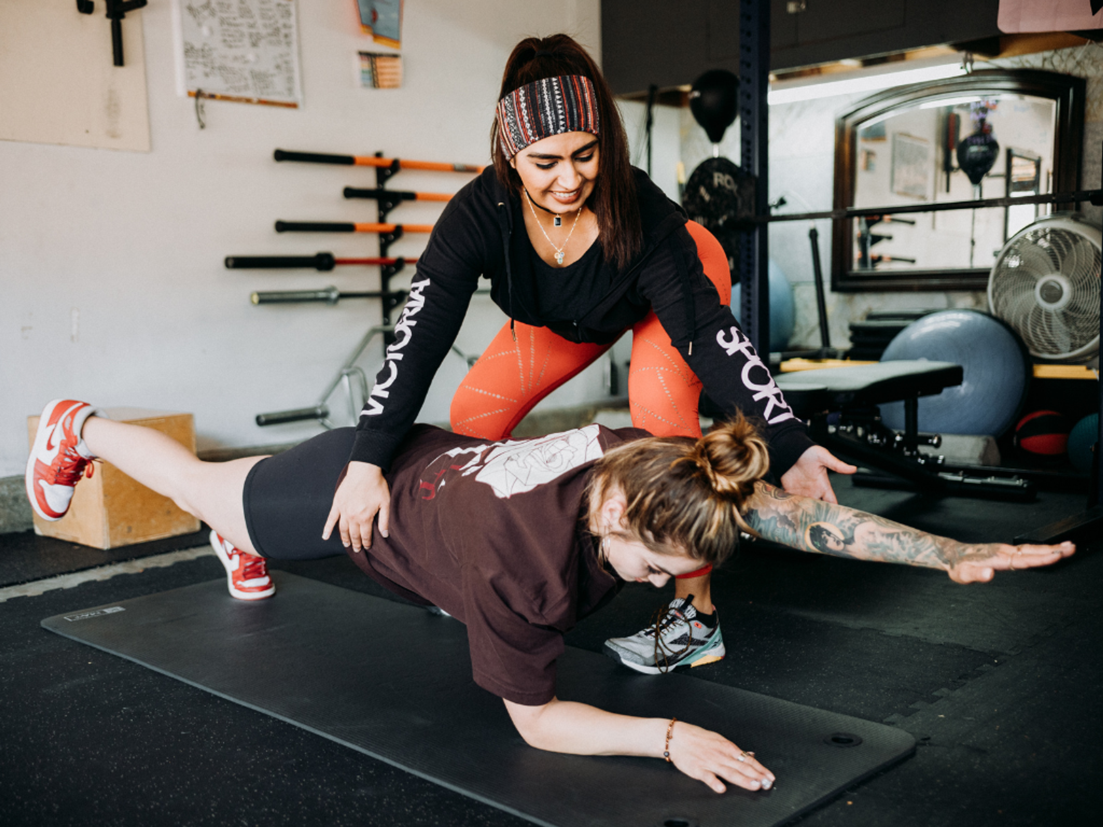 A fitness instructor assists a woman performing a plank with one arm and leg extended in a gym.