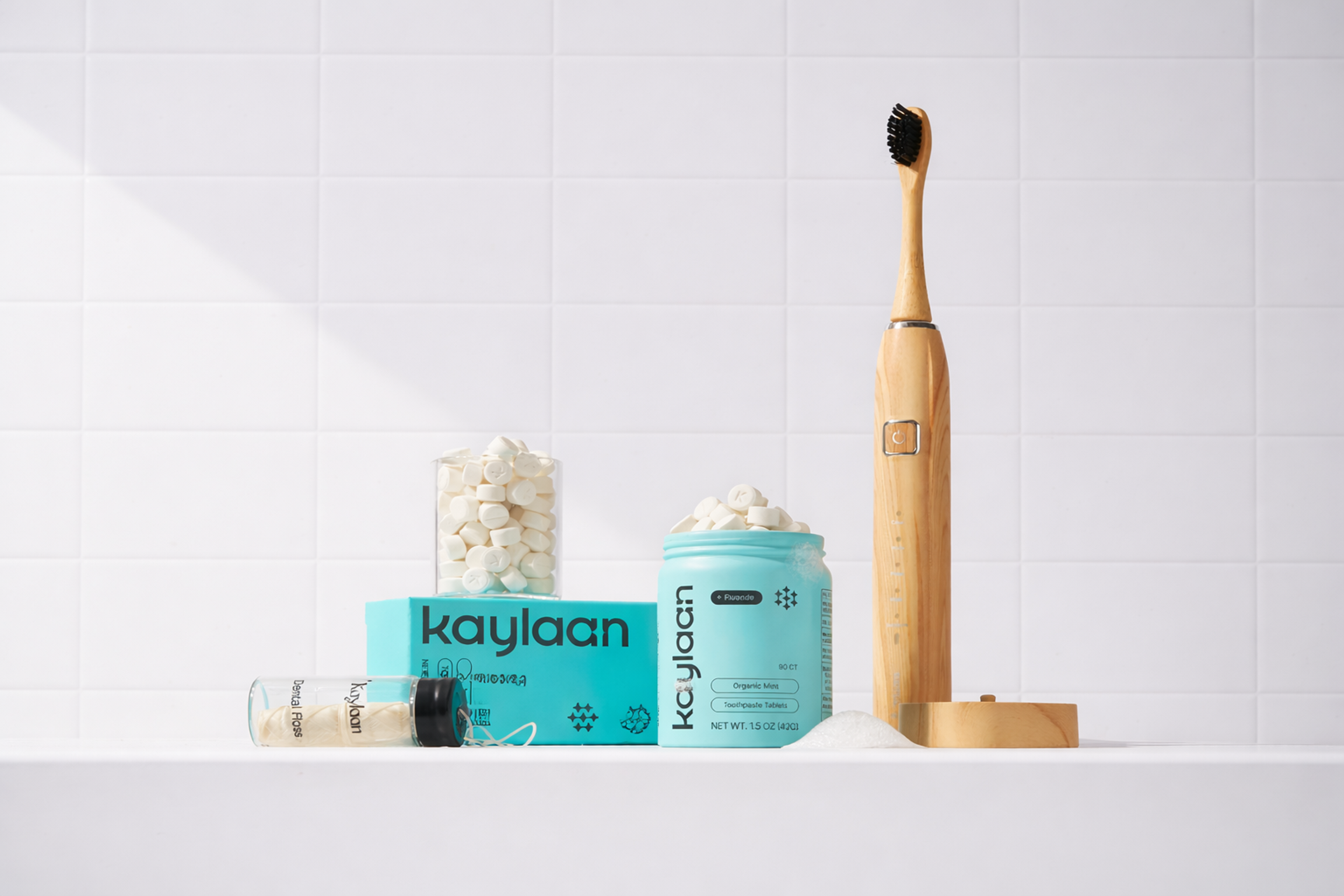 Kaylaan dental products: a wooden electric toothbrush, turquoise jar of toothpaste tablets, and dental floss, on a white tiled surface.