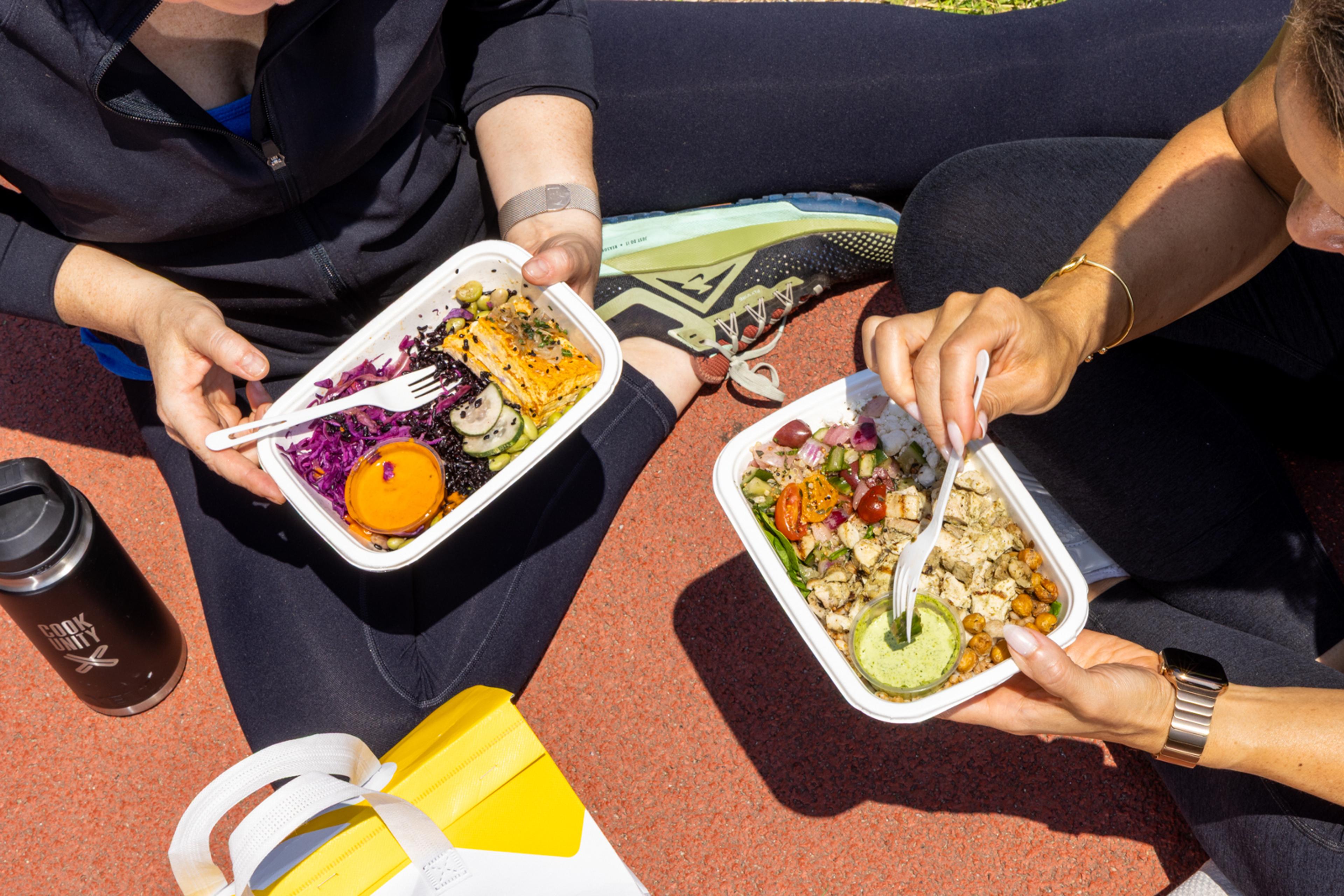 Two people in athletic wear eating CookUnity meal bowls on a running track