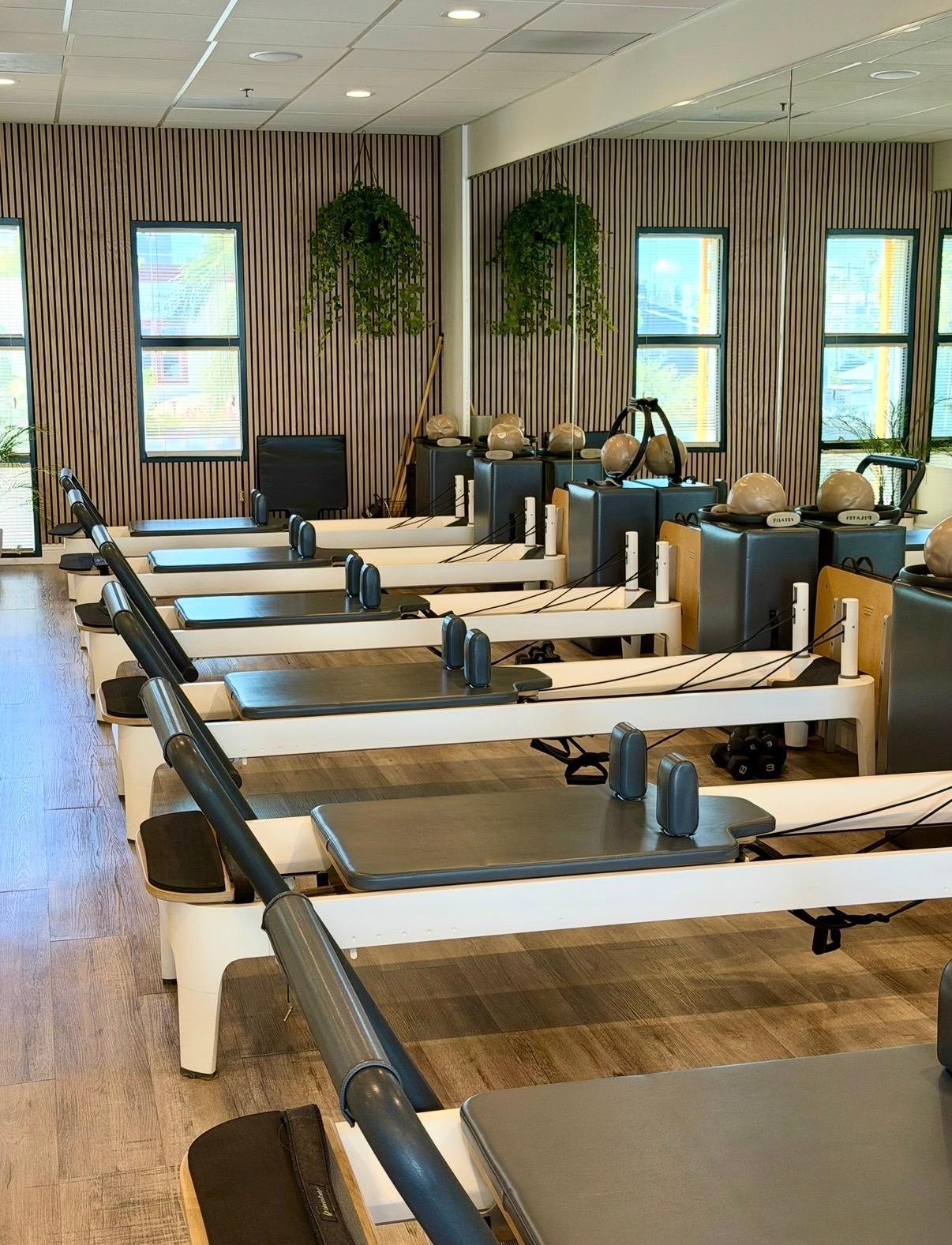 A bright pilates studio with several reformers, wood-slatted walls, and hanging plants.