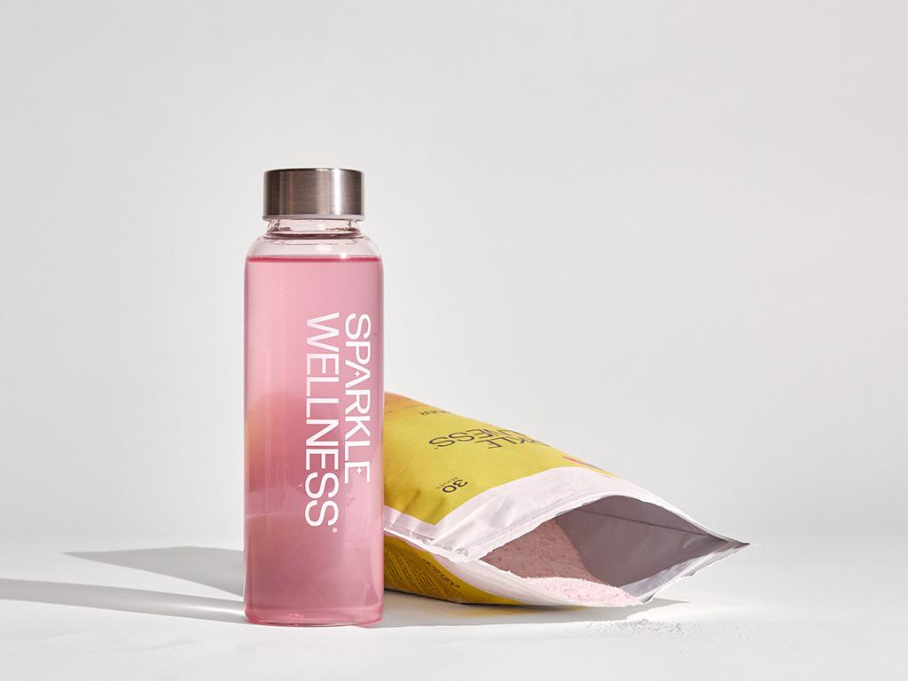 A clear bottle of pink "Sparkle Wellness" liquid next to an open yellow pouch with pink powder.