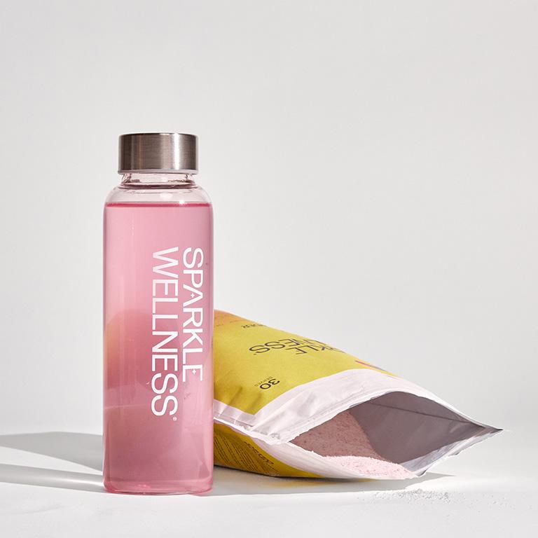 A clear bottle of pink "Sparkle Wellness" liquid next to an open yellow pouch with pink powder.