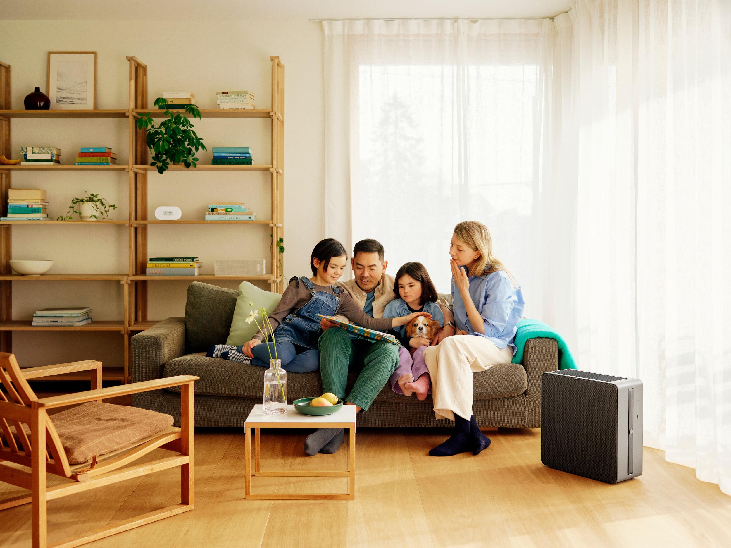 A family and their dog relax on a sofa, looking at a book, with an air purifier visible in their bright living room.
