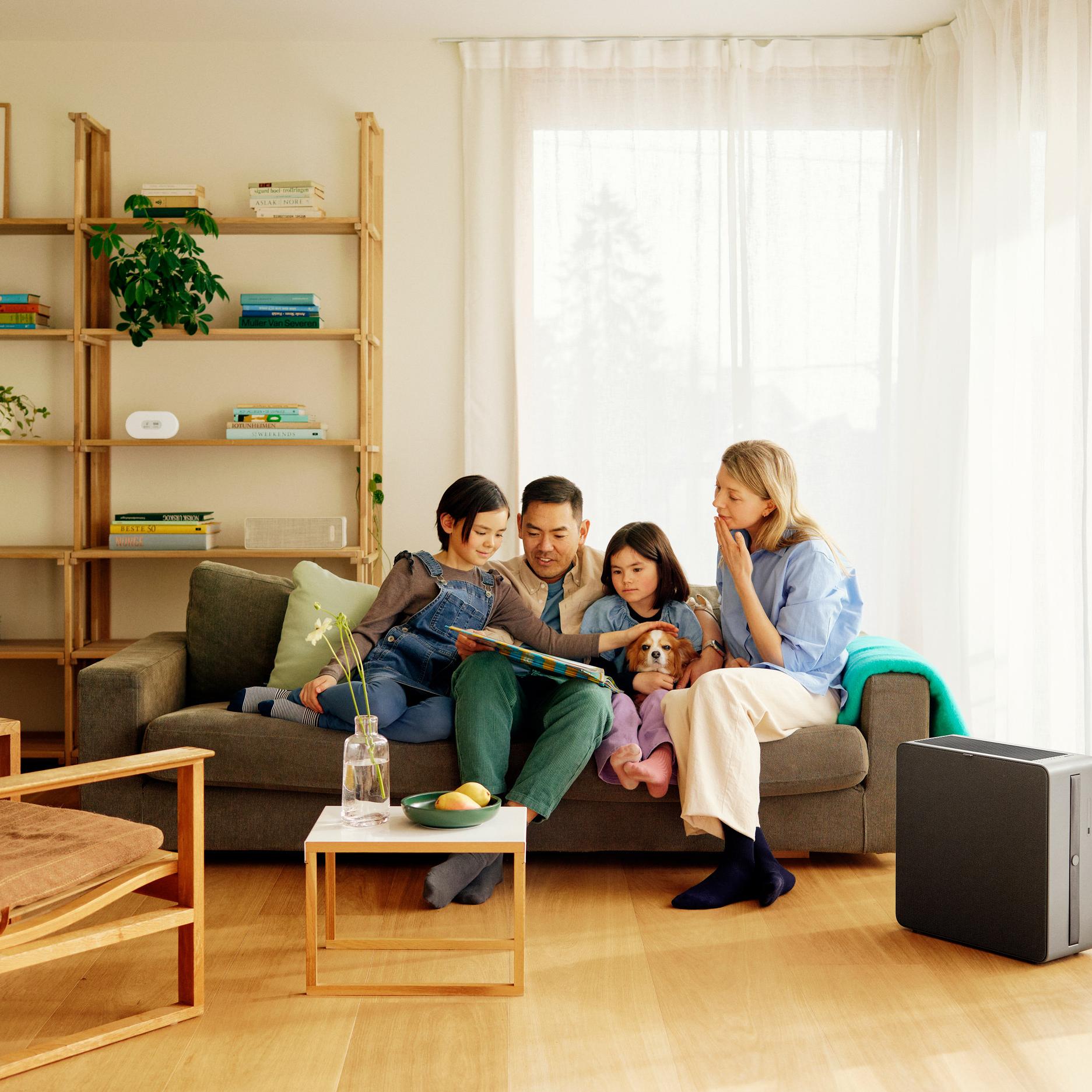 A family and their dog relax on a sofa, looking at a book, with an air purifier visible in their bright living room.