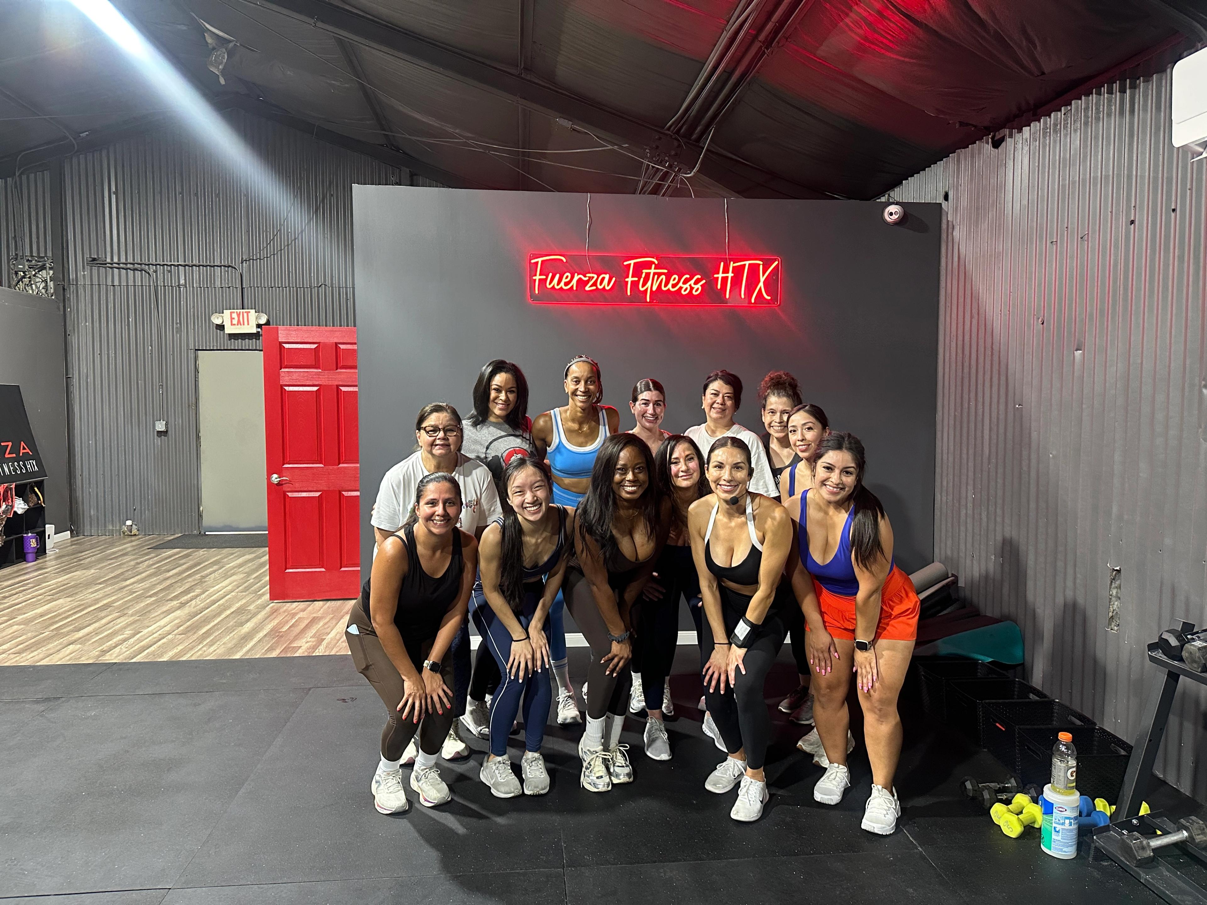 A diverse group of women in athletic wear posing and smiling in a gym with a 'Fuerza Fitness HTX' neon sign.