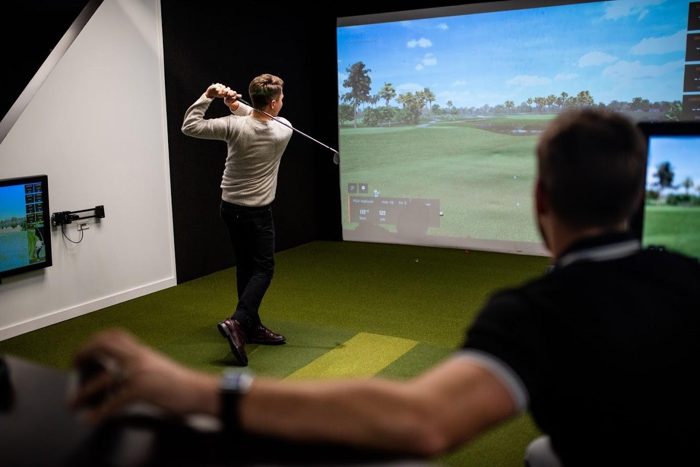 A man swings a golf club in an indoor golf simulator, aiming at a large screen displaying a virtual golf course.