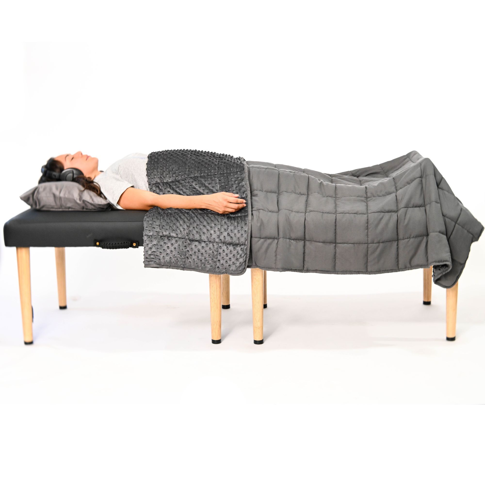 A woman wearing headphones lies on a massage table, covered by a grey textured blanket and a grey weighted blanket.