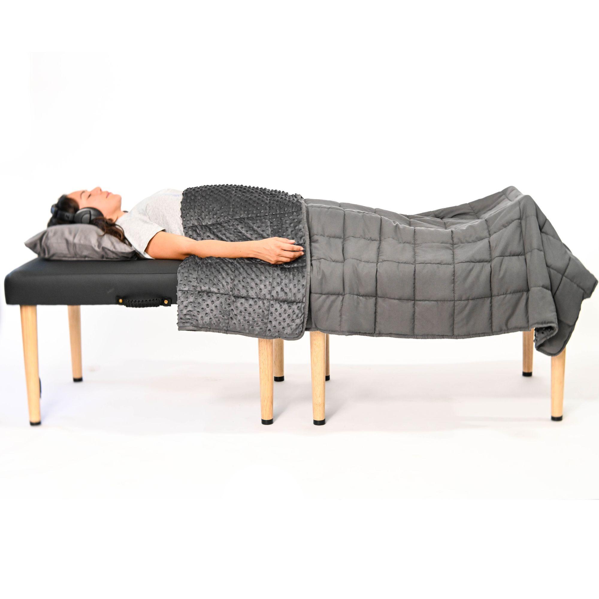 A woman wearing headphones lies on a massage table, covered by a grey textured blanket and a grey weighted blanket.