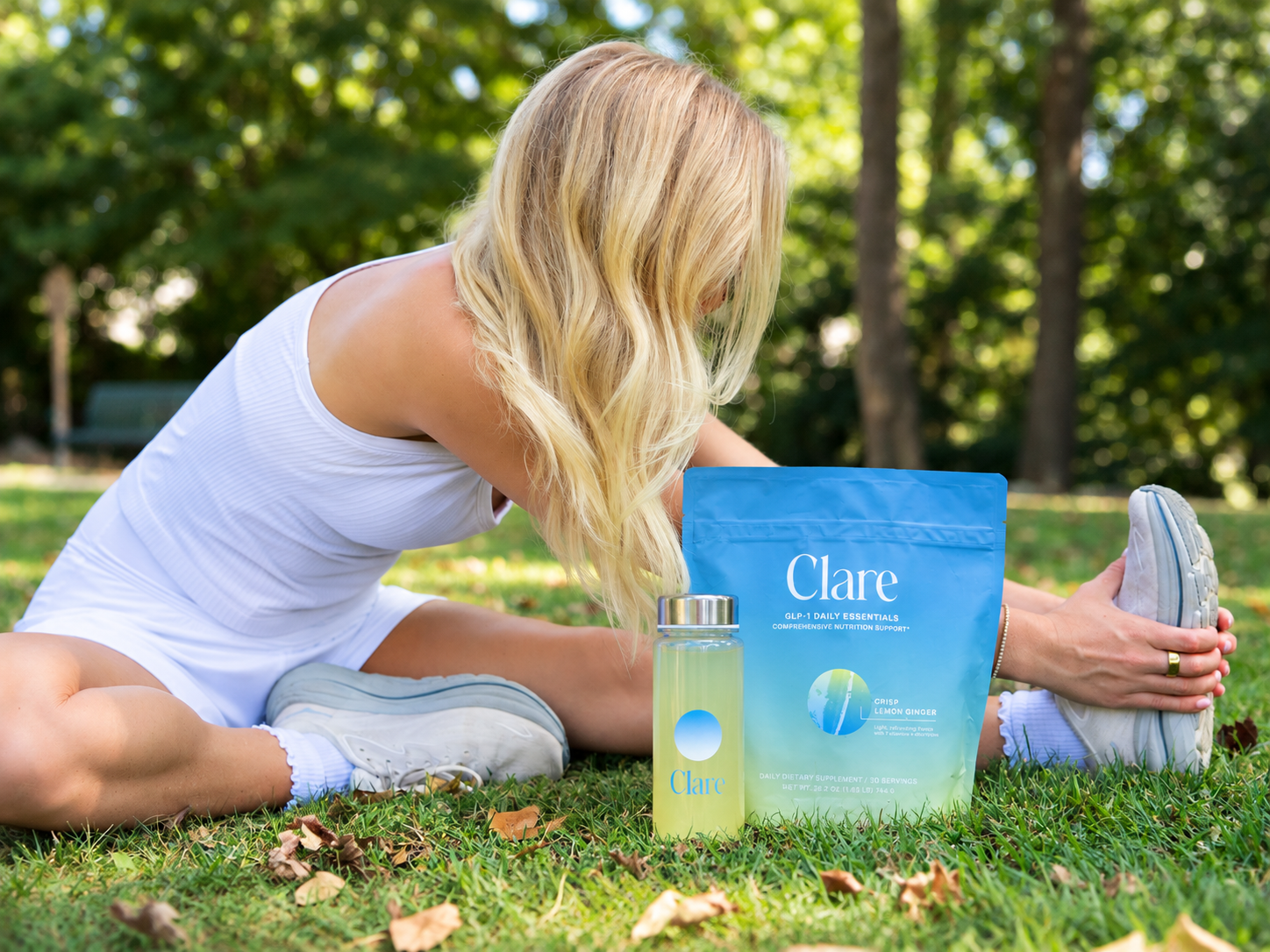 A blonde woman in a white athletic dress stretches on grass next to a Clare branded drink bottle and supplement pouch.