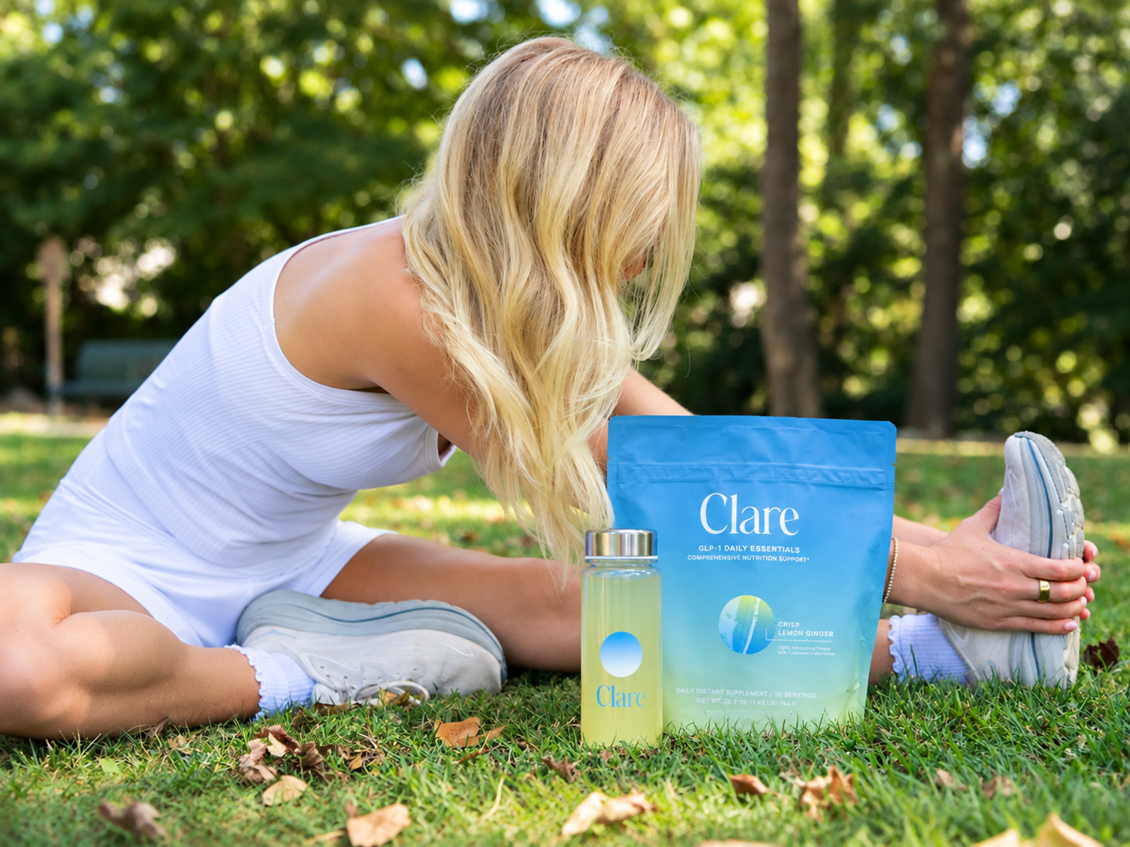 A blonde woman in a white athletic dress stretches on grass next to a Clare branded drink bottle and supplement pouch.
