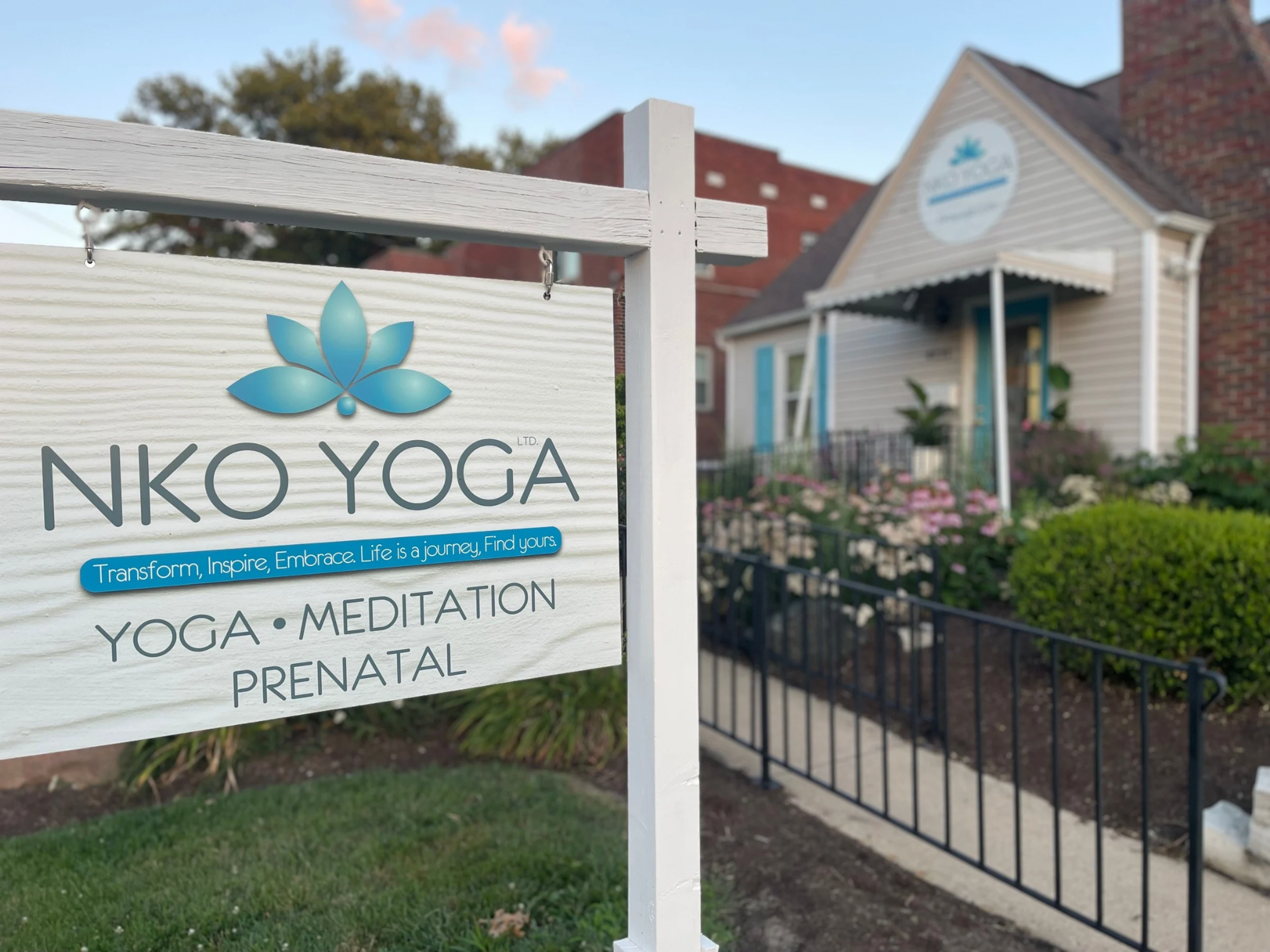 A white sign for NKO YOGA, featuring a blue lotus logo and listing yoga, meditation, and prenatal services, stands outside a cream-colored studio building with blue accents.