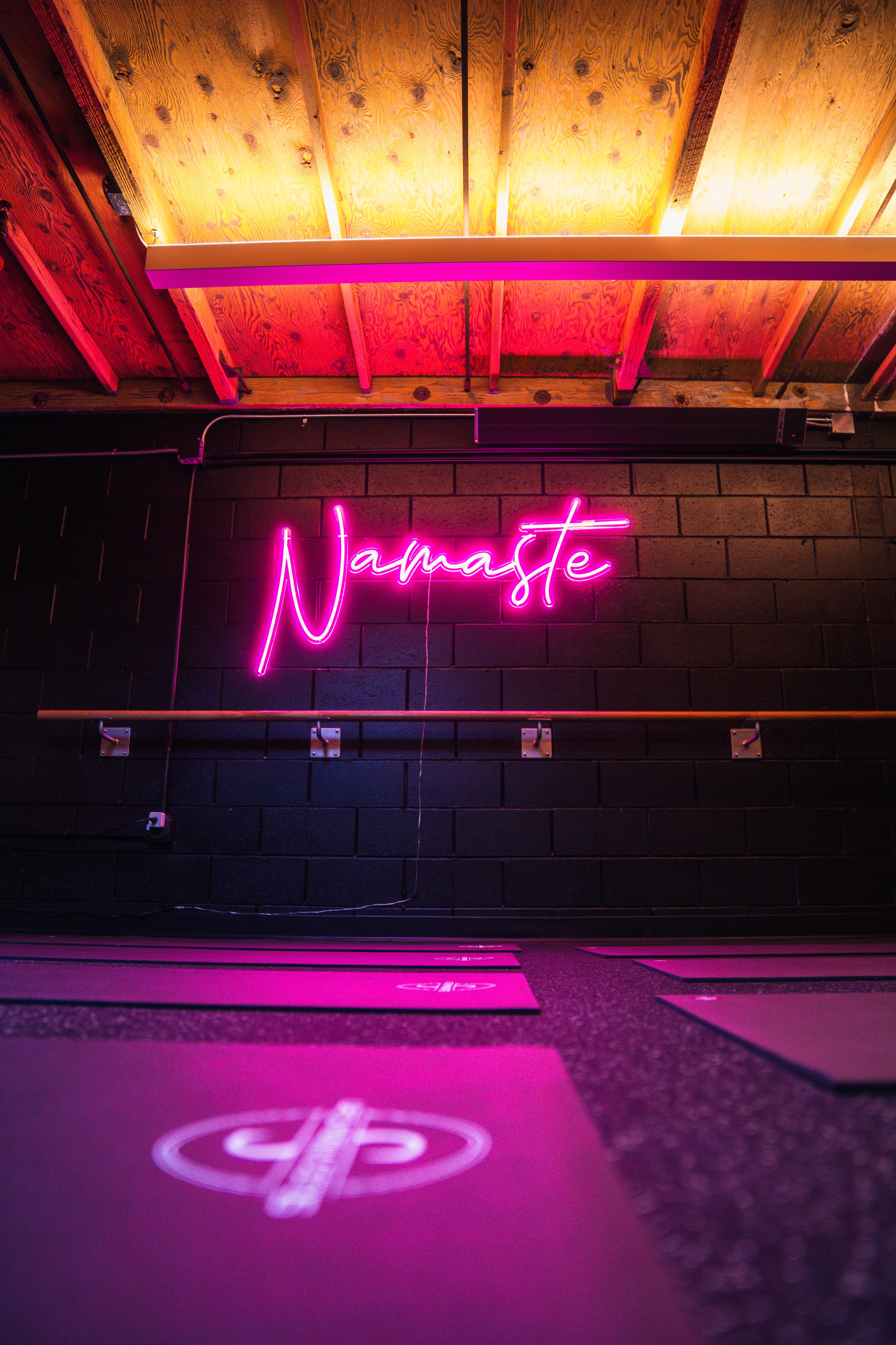 A pink neon "Namaste" sign on a dark brick wall, with a barre below and several yoga mats on the floor illuminated by a pink glow.