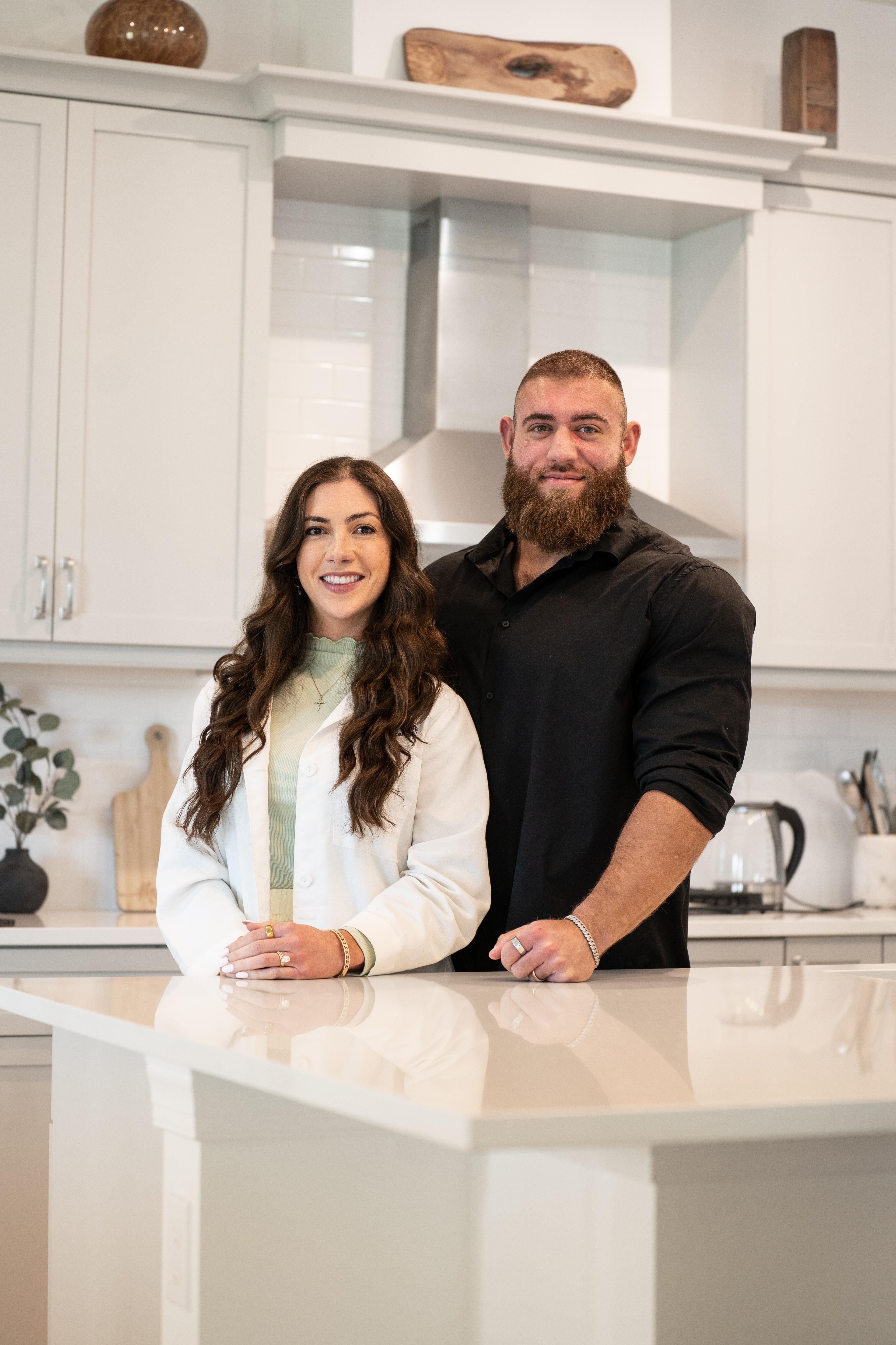 A smiling man with a beard and a smiling woman stand in a modern kitchen.