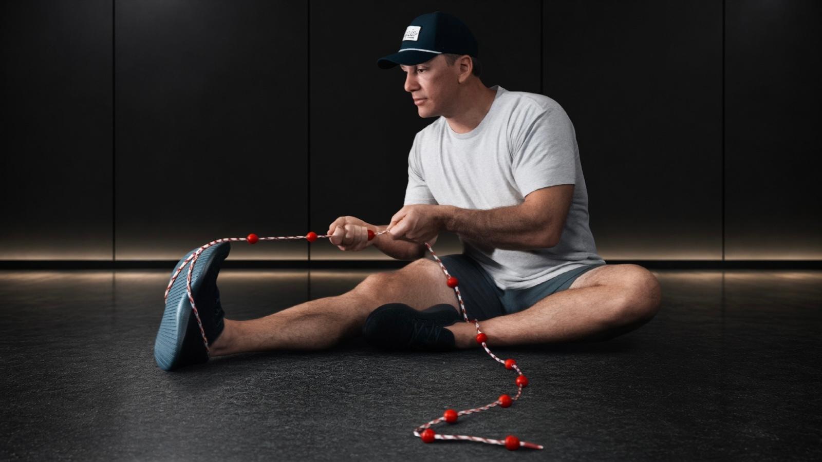 Man sits, using a red and white beaded rope for a foot exercise.