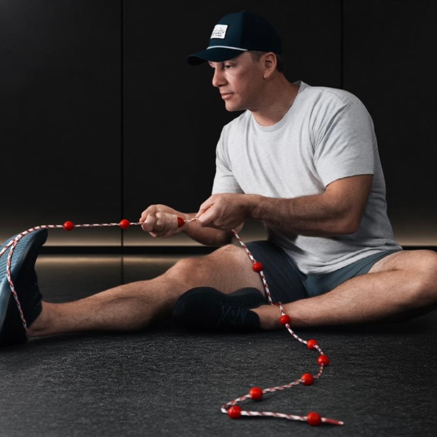 Man sits, using a red and white beaded rope for a foot exercise.