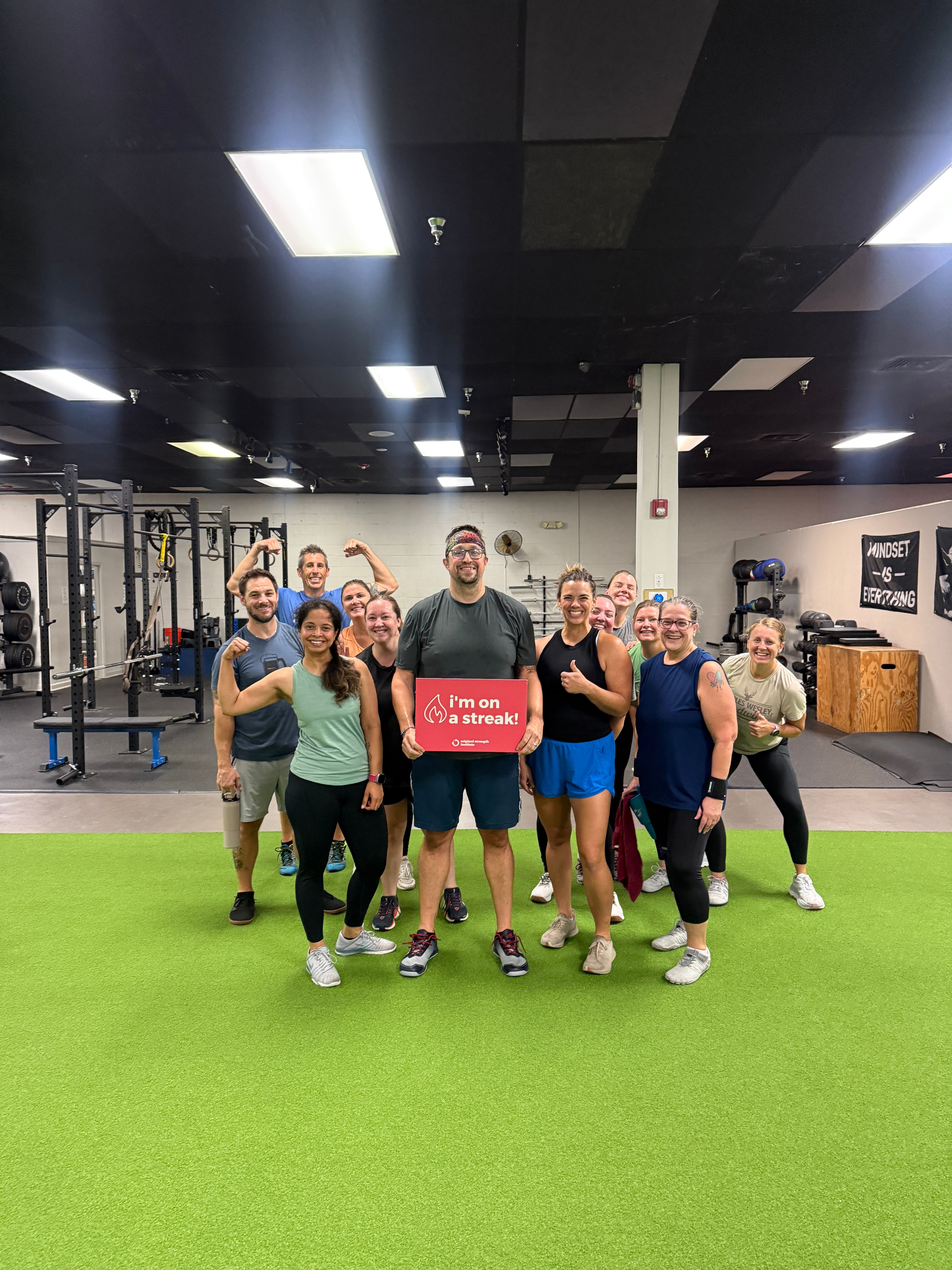 A group of people smiling and posing in a gym, one person holding a red sign that reads "I'm on a streak!".