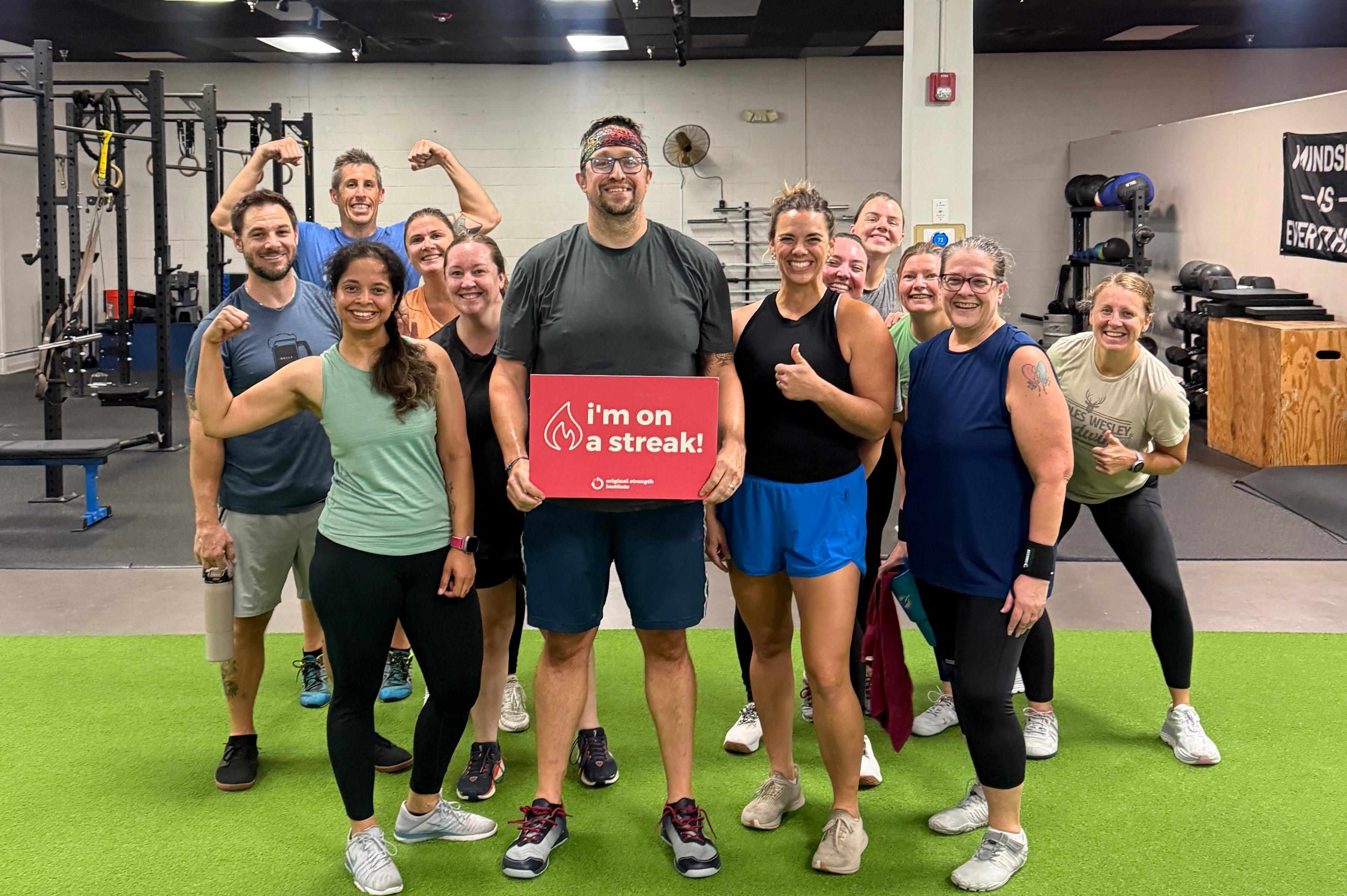 A group of people smiling and posing in a gym, one person holding a red sign that reads "I'm on a streak!".